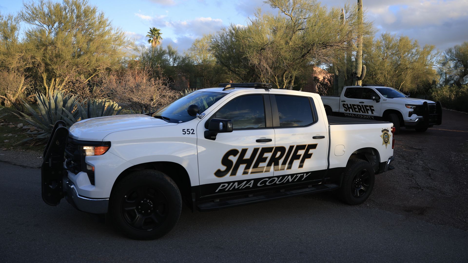Two white Pima County sheriff pickup trucks parked near desert vegetation including cacti and trees under a partly cloudy blue sky.