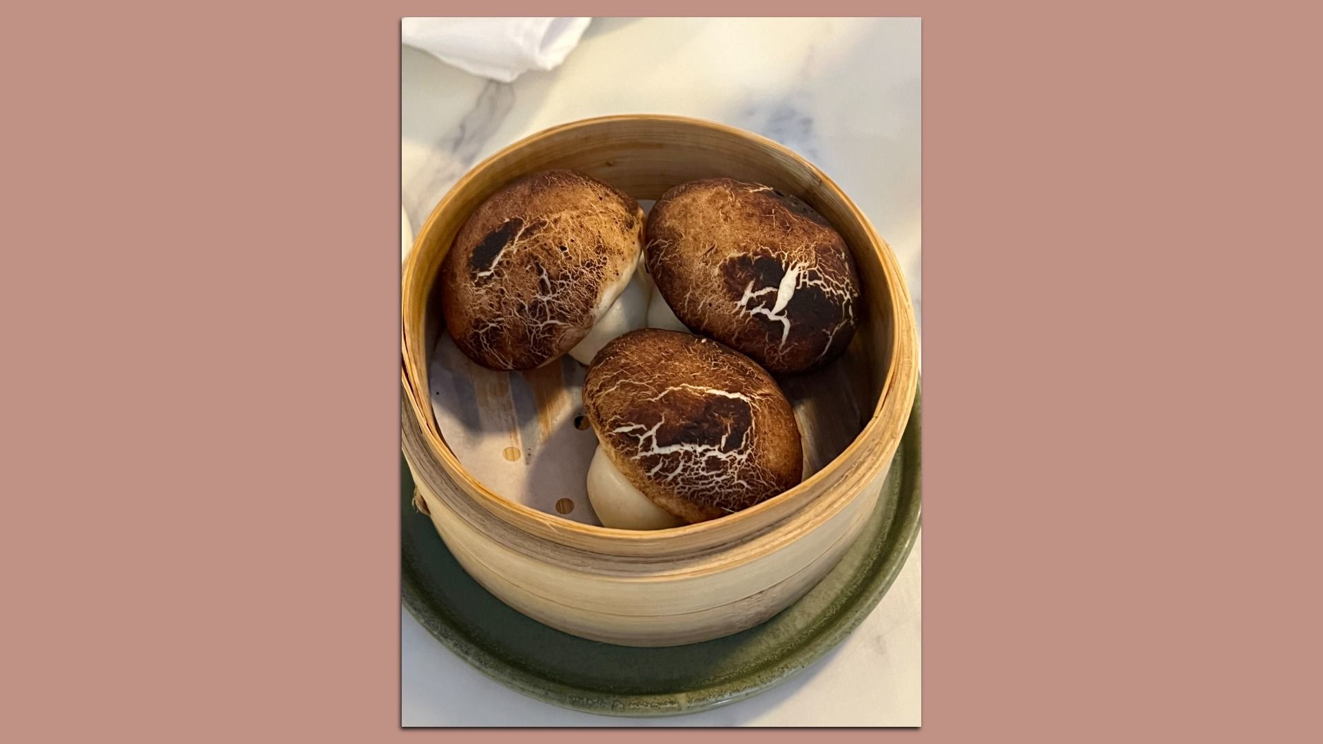 Three steamed buns shaped and colored like brown mushrooms with white veiny patterns, served in a round bamboo steamer on a green plate.