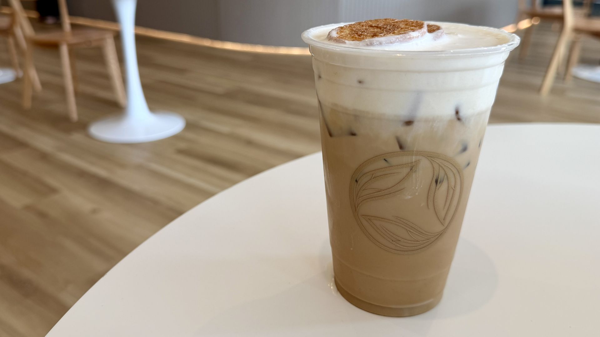 Iced coffee layered with milk and ice, topped with whipped cream and a caramel disc, in a plastic cup on a white table; blurred café interior with wooden floor in the background.