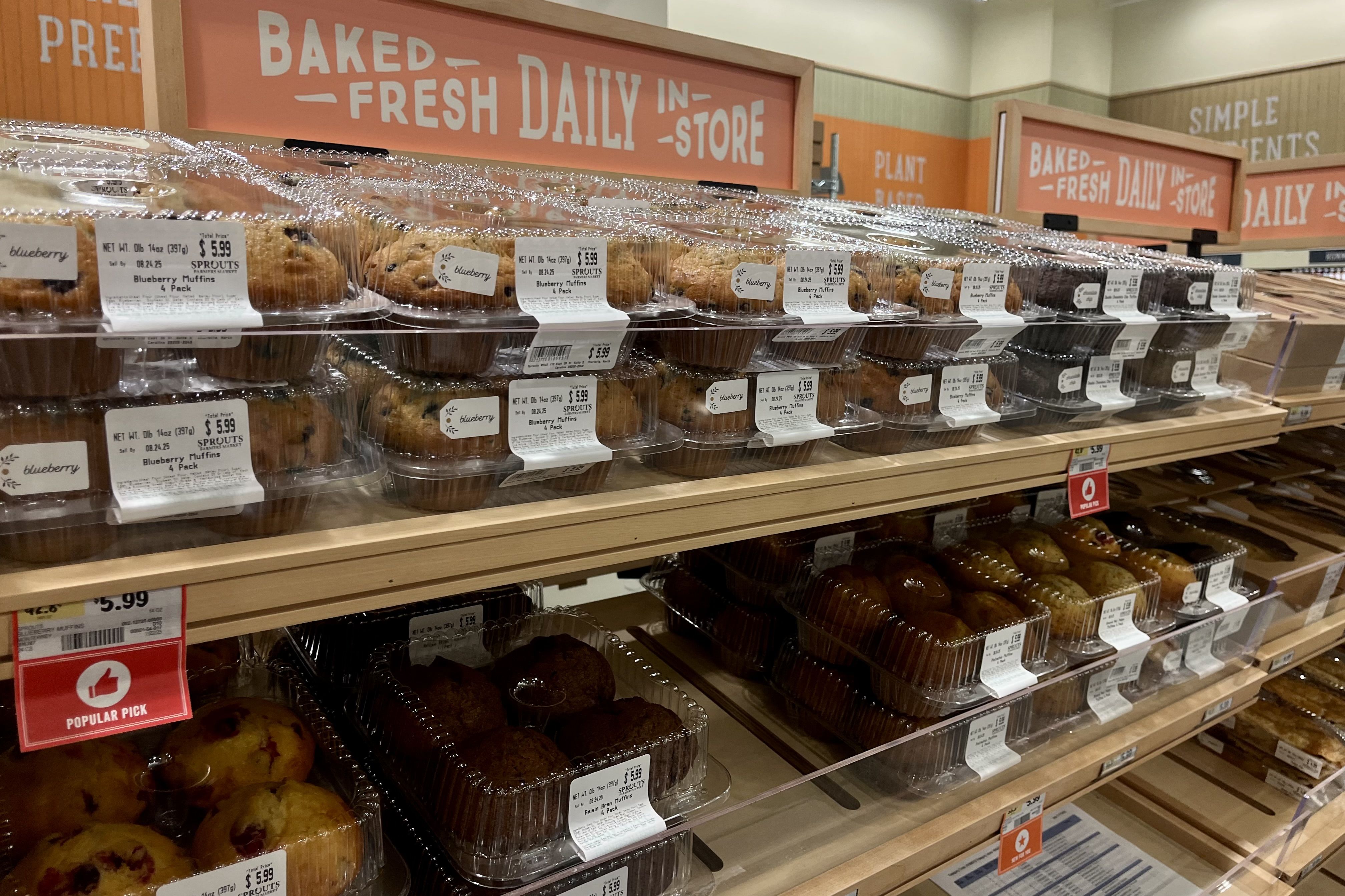 Shelves in a store bakery section displaying various packaged muffins, including blueberry and other flavors, with signs reading "Baked Fresh Daily in Store" and price tags visible.