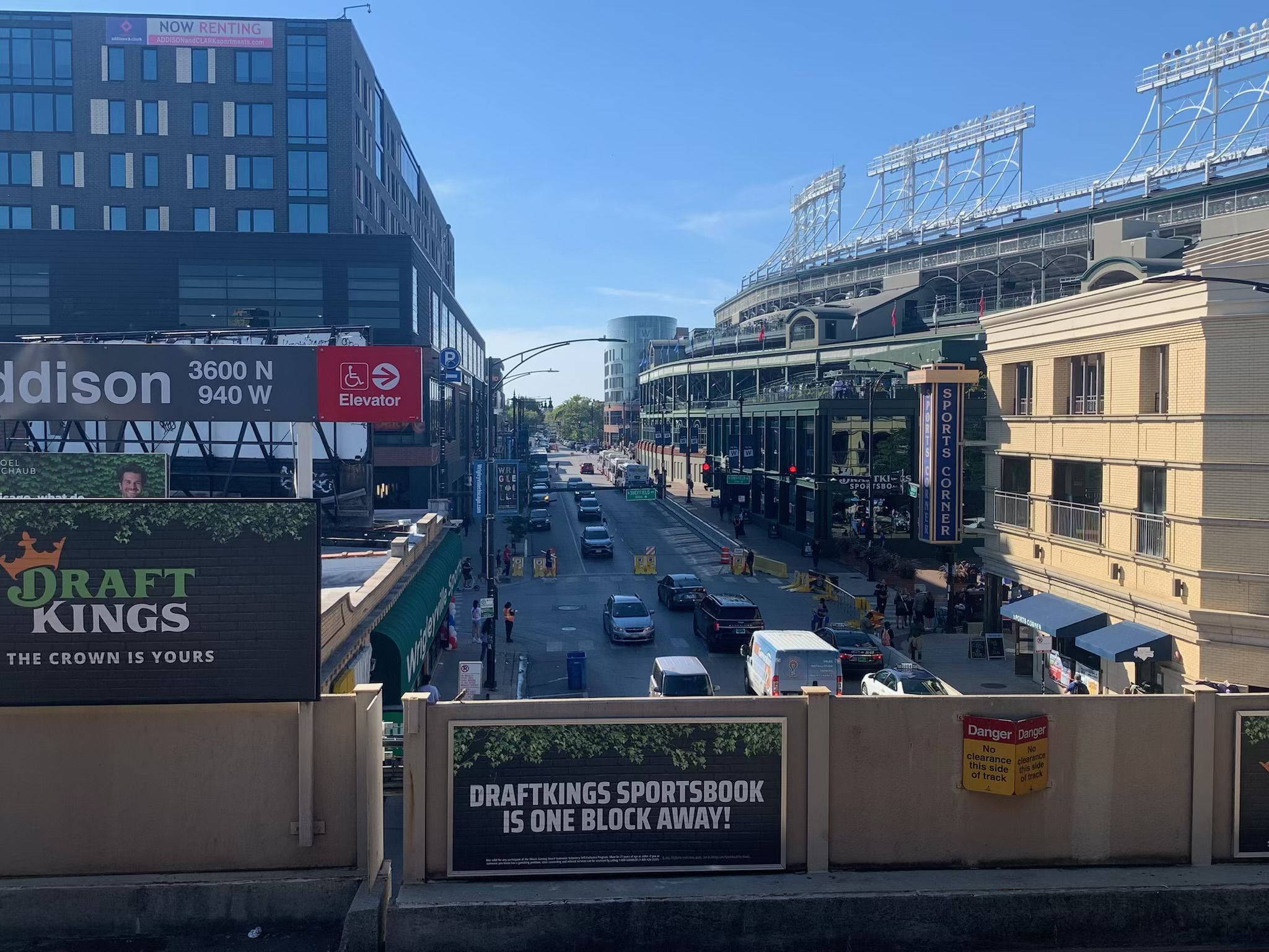 Street view of a Clark Street on a sunny day with traffic, signs for Addison station, DraftKings ads, and Wrigley Field sports corner visible in the background.