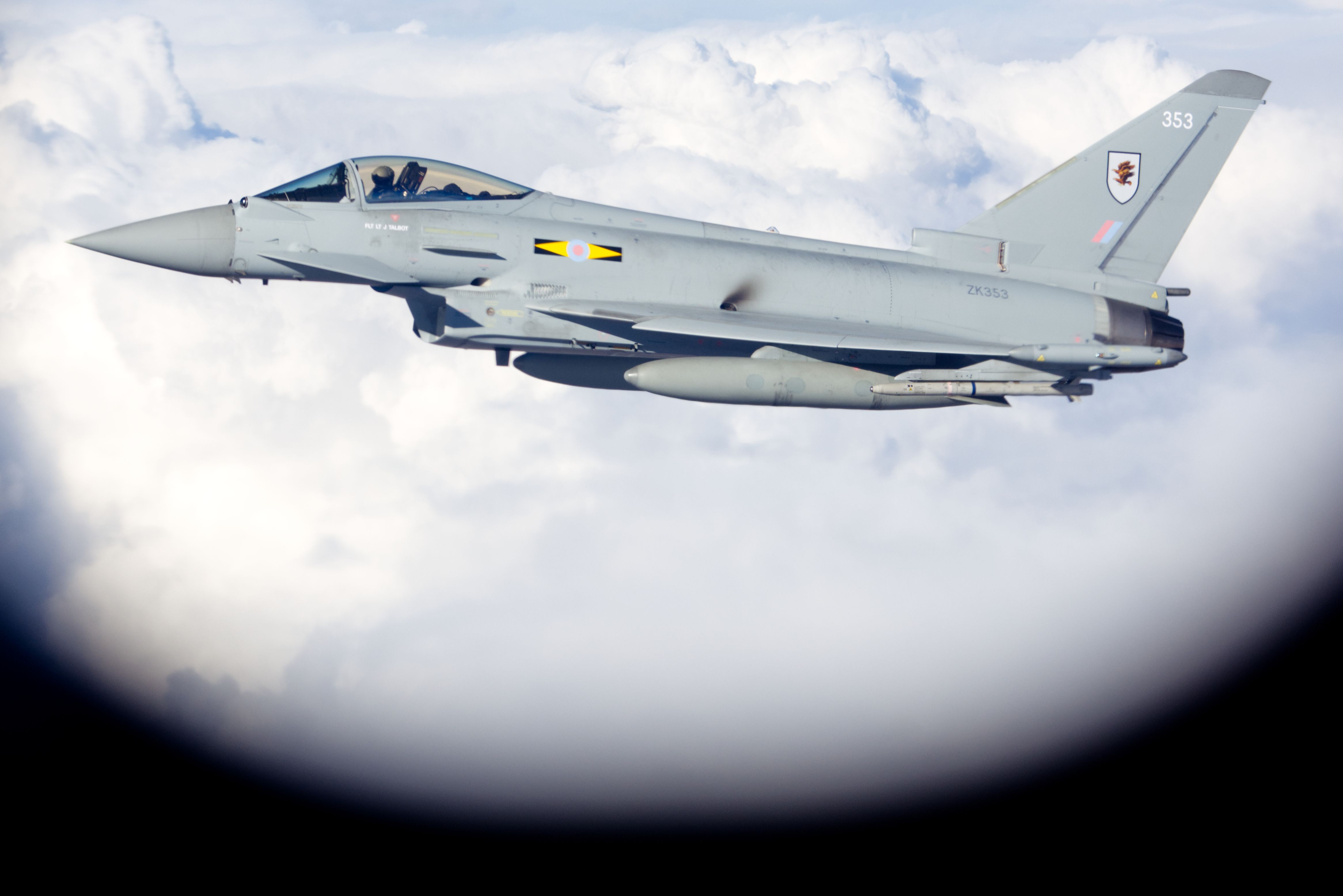 Gray military fighter jet flying above clouds, marked with number 353 and a winged emblem on the tail fin, viewed through a circular frame against a bright sky.