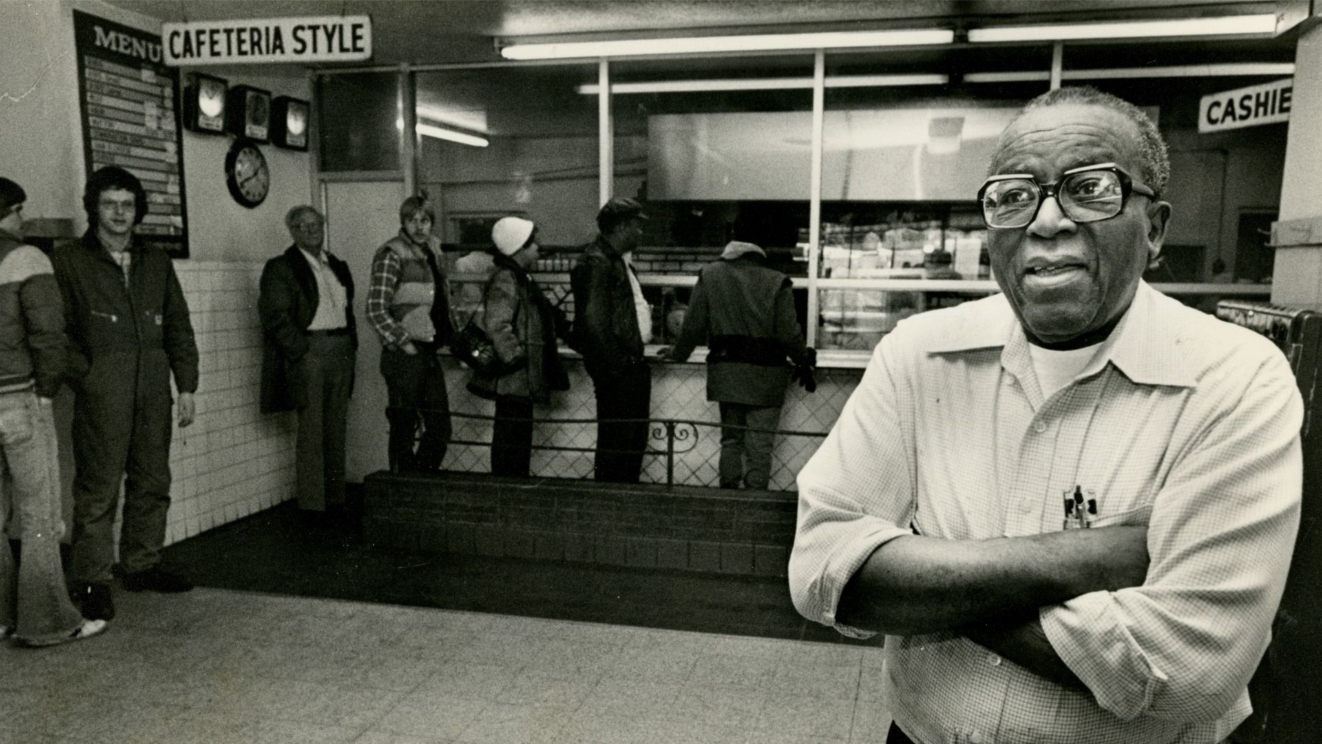 Black and white photo of a man with glasses and folded arms in the foreground of a cafeteria. People queue up at the serving counter under a "CAFETERIA STYLE" sign.