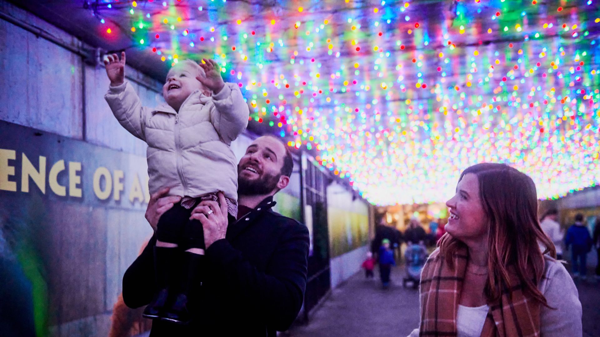 Man lifting a smiling child in a white coat reaching towards colorful overhead lights, with a woman in a plaid scarf smiling at them in a festive tunnel.