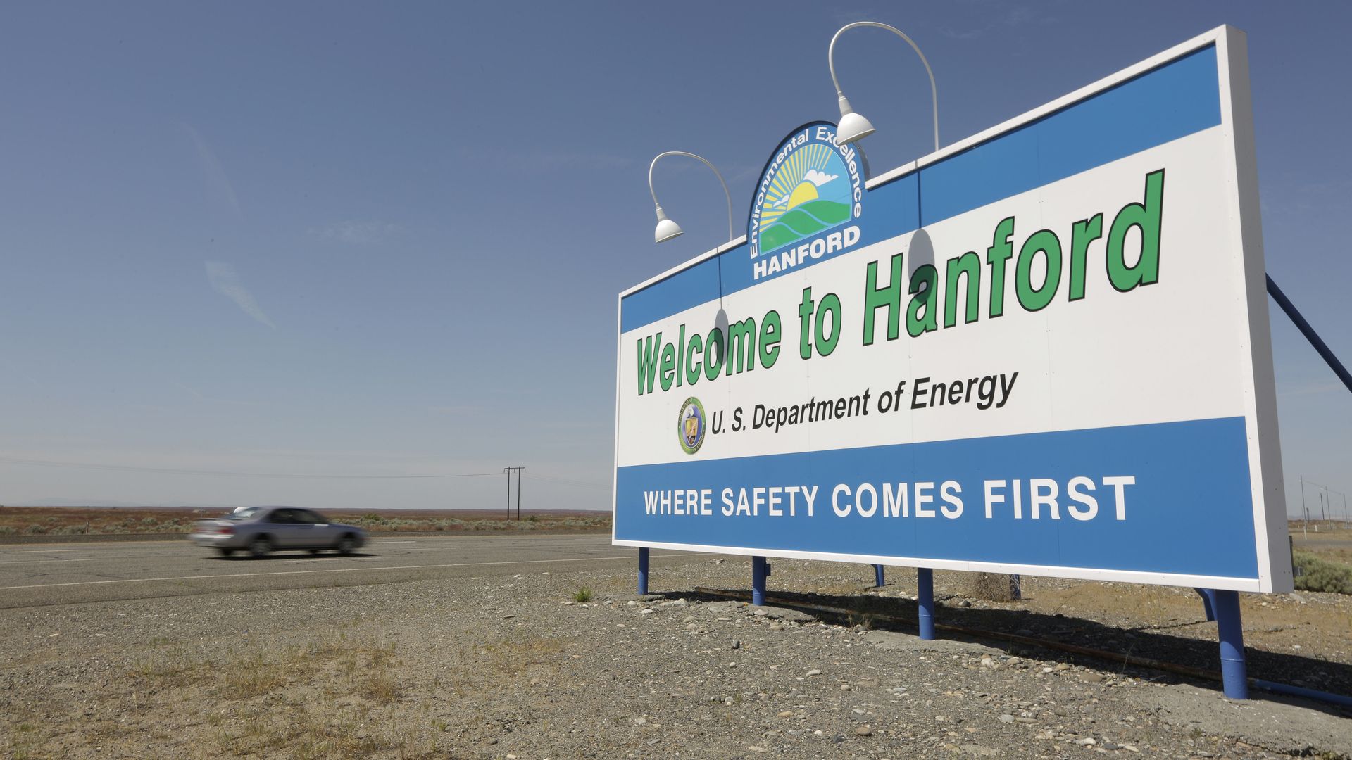 Large roadside sign reading "Welcome to Hanford" with logo and "U.S. Department of Energy" in green, black, and blue text, set against a clear sky and dry landscape with a passing car.