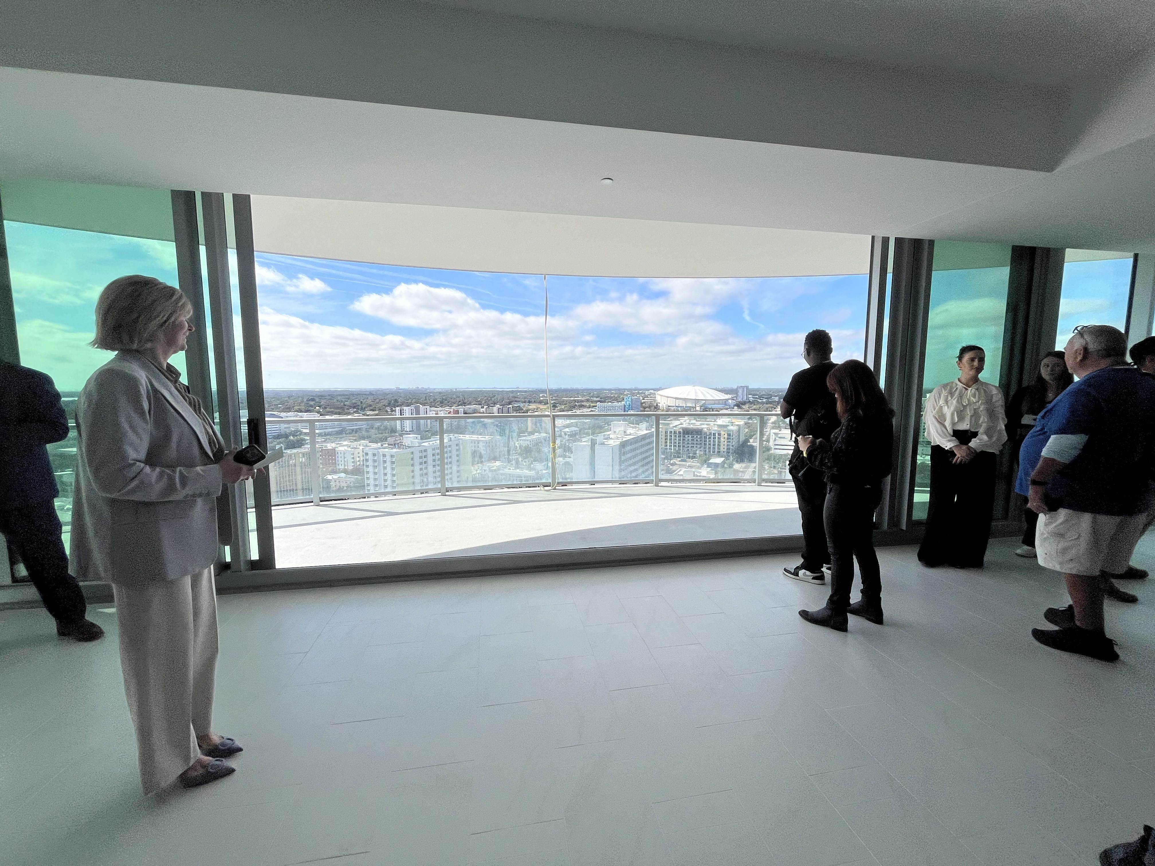 Group of people inside a modern room with large floor-to-ceiling glass windows overlooking a cityscape under blue sky with clouds, featuring a dome-shaped building in the distance.