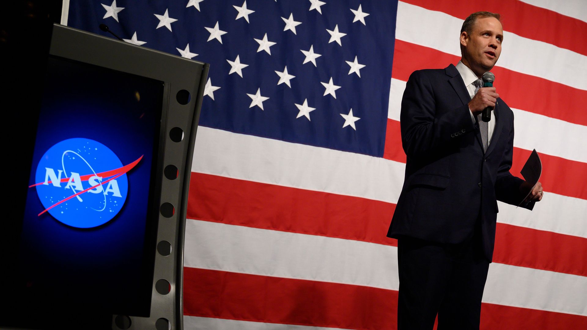 In this image, a man in a suit holds and speaks into a microphone with the American flag behind him.