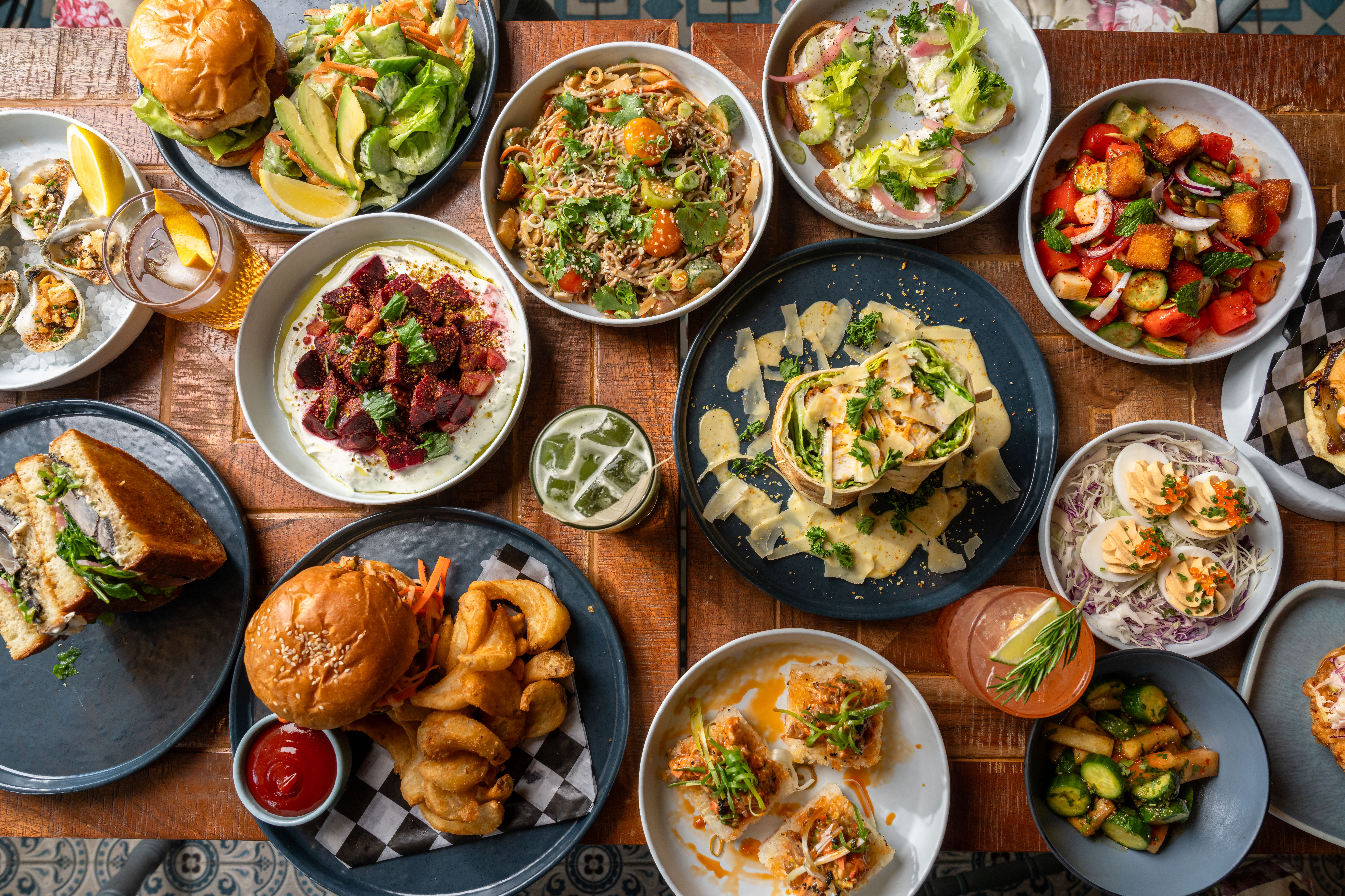 Top-down view of a wooden table crowded with a colorful feast: burgers, sandwiches, fresh salads with avocado, a central noodle pasta bowl, tacos, roasted vegetables, and drinks.