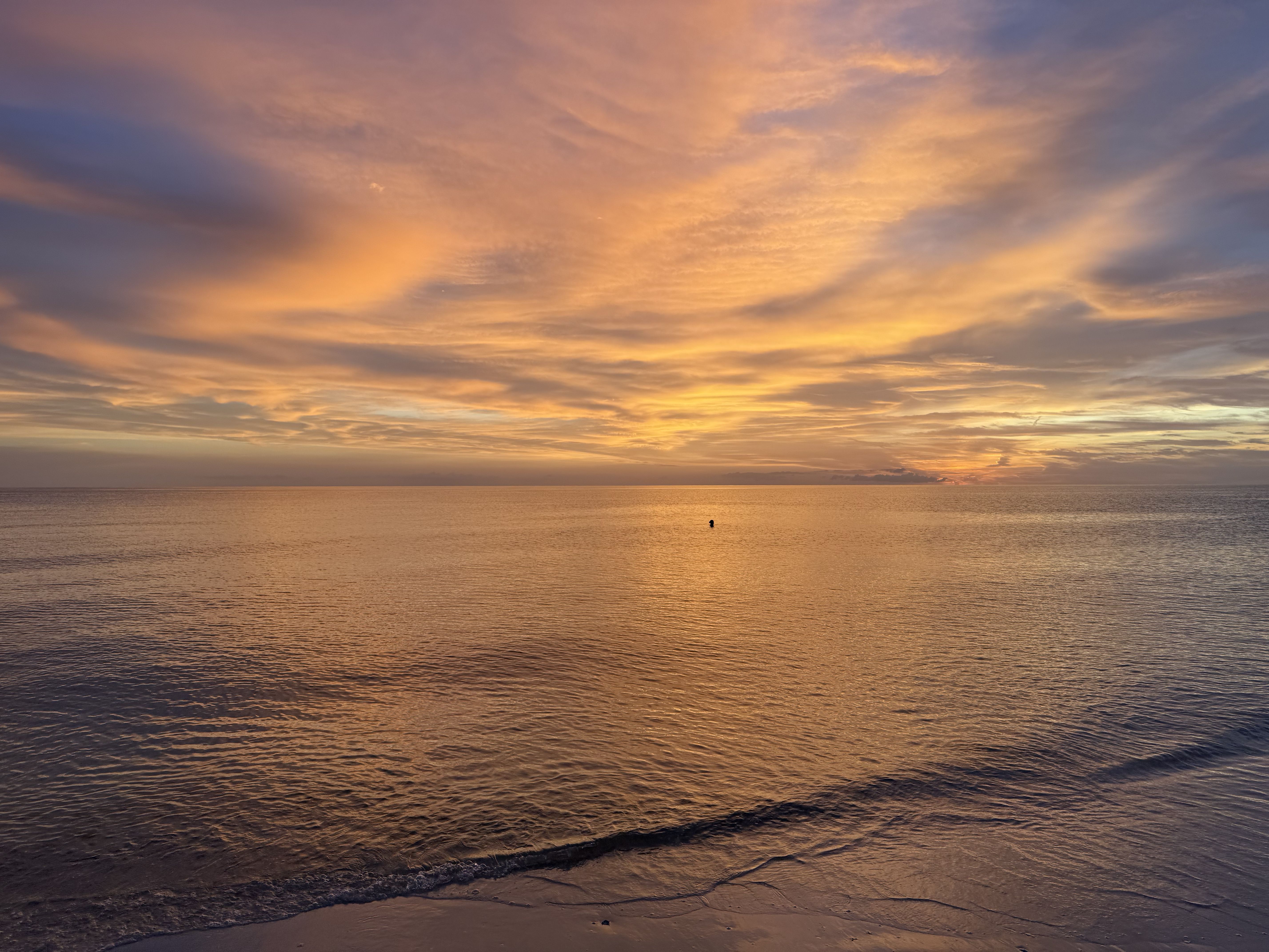 Calm ocean at sunset with vibrant orange, yellow, and purple clouds reflecting on smooth water, gentle waves touching sandy shore under a partly cloudy sky.