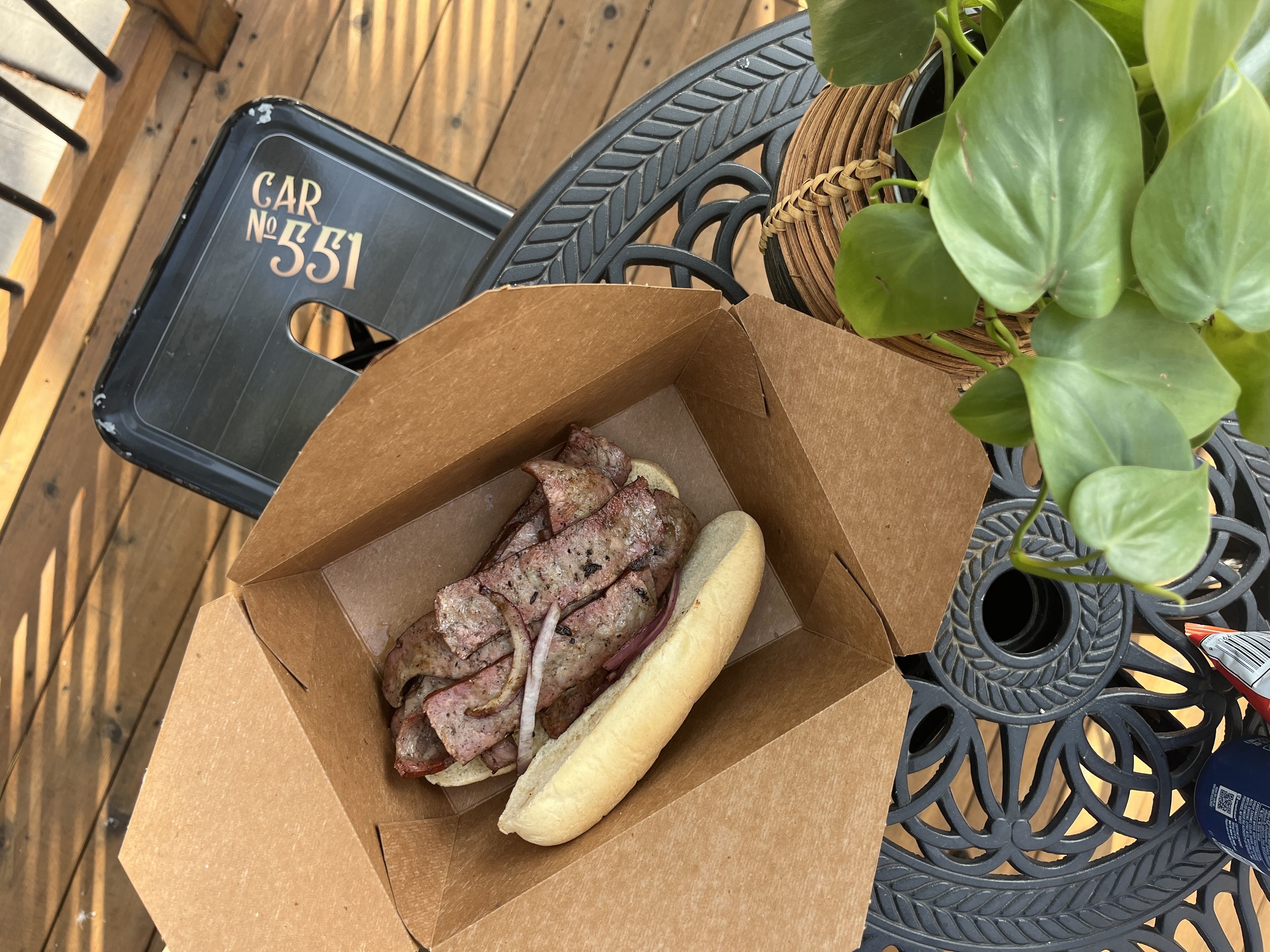Grilled sausage sandwich with onions in a brown cardboard takeout box on a black metal table with a green potted plant nearby, on a wooden deck with a black chair labeled "Car No 551".