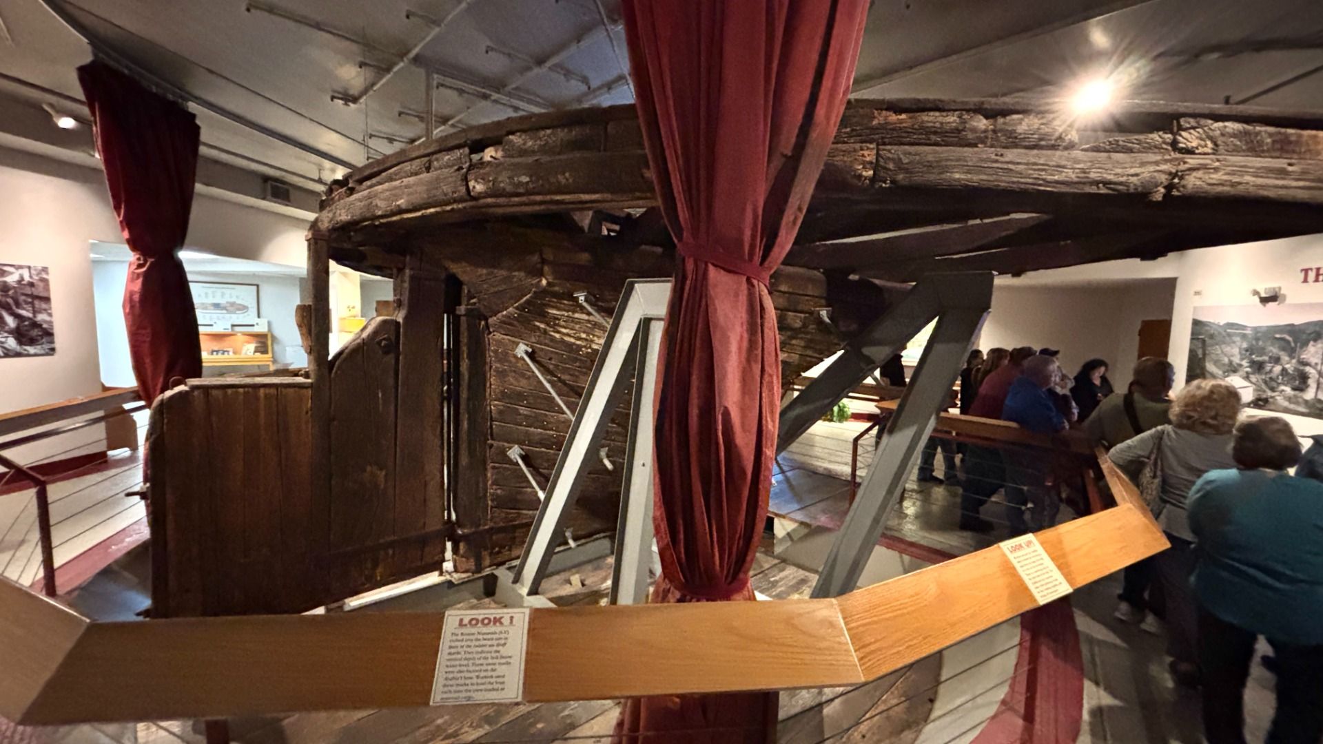 Indoor museum exhibit with a large, dark wooden ship hull overhead a circular display. Red curtains frame the area; visitors sit on benches to the right, with wall displays in the background.