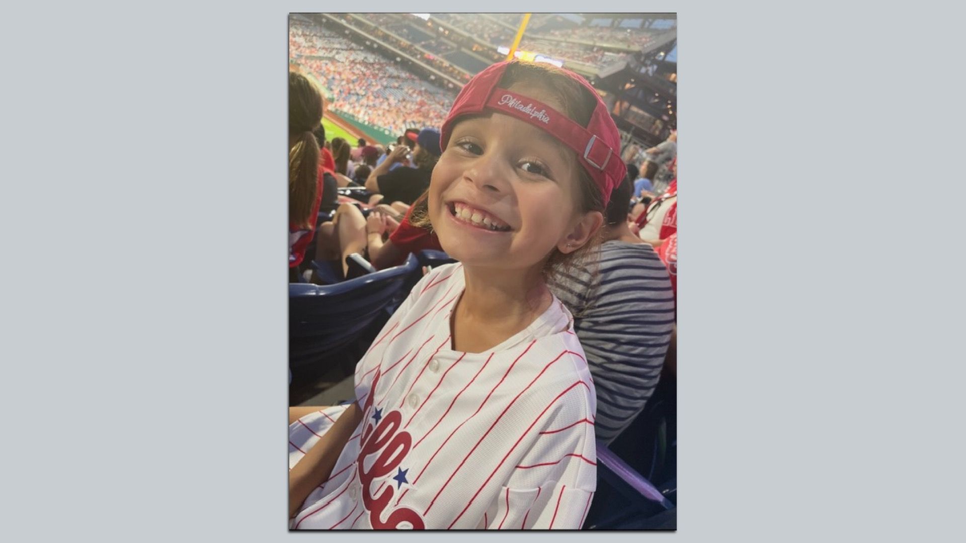 Philly elementary student Isabella Gonzalez wearing a red Phillies cap backward and a white pinstripe jersey, smiling at a baseball stadium with fans in the stands.