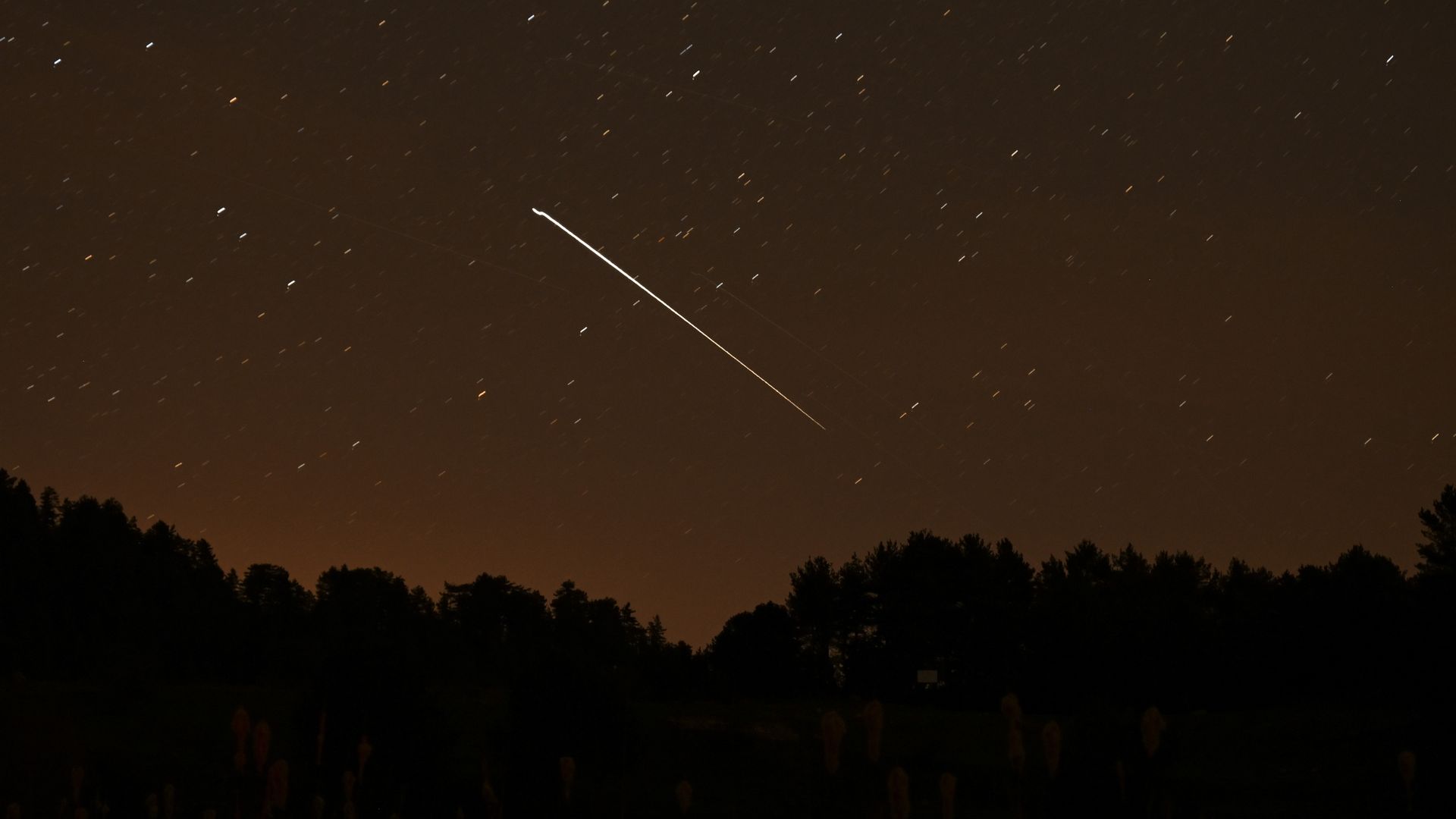 Long-exposure night scene: orange-tinted horizon, silhouetted trees and grasses in the foreground, with numerous star trails and a bright diagonal meteor-like streak across the sky.
