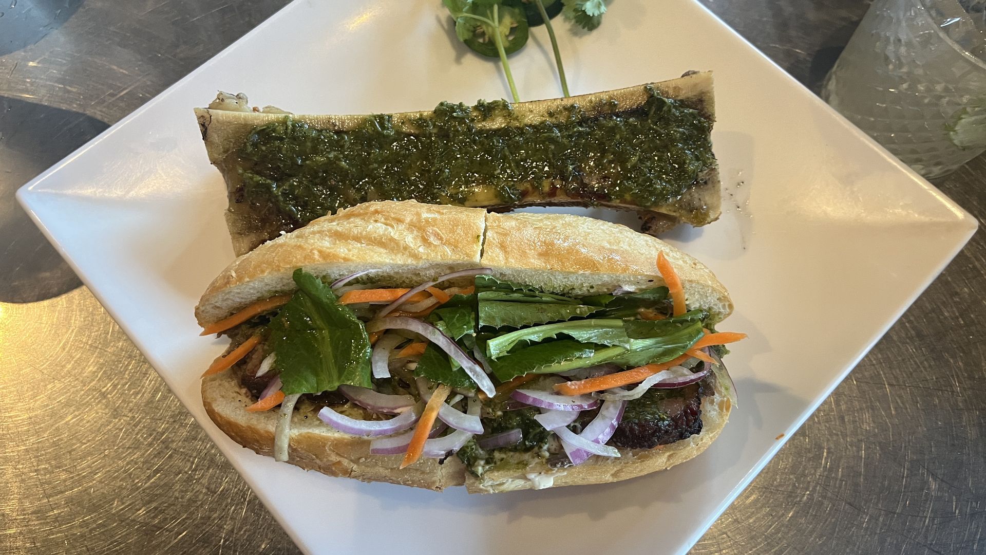 A banh mi topped with carrots, red onion and lettuce sits on a plate next to a bone marrow topped with chimichurri and cilantro and jalapeños on the side.