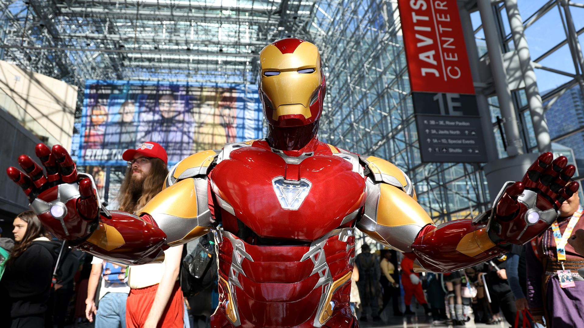 Iron Man statue in red and gold stands with arms raised in a glass atrium, surrounded by people; a red Javits Center banner and steel beams loom in the bright background.