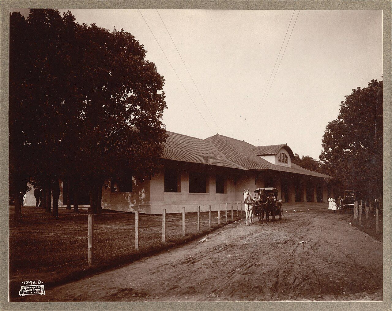 Vintage sepia photo of a single-story train station with a tiled roof, row of windows, a horse-drawn carriage on a dirt road in front, and tall trees framing the scene.