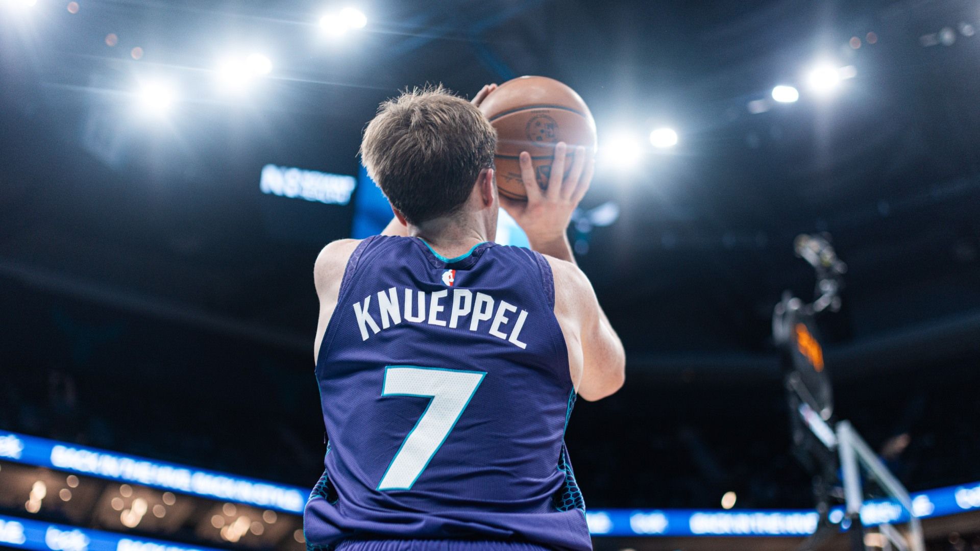 Basketball player seen from behind, wearing a purple jersey with the name KNEEPPEL and the number 7, holding a ball at chest level in a packed arena under bright lights.