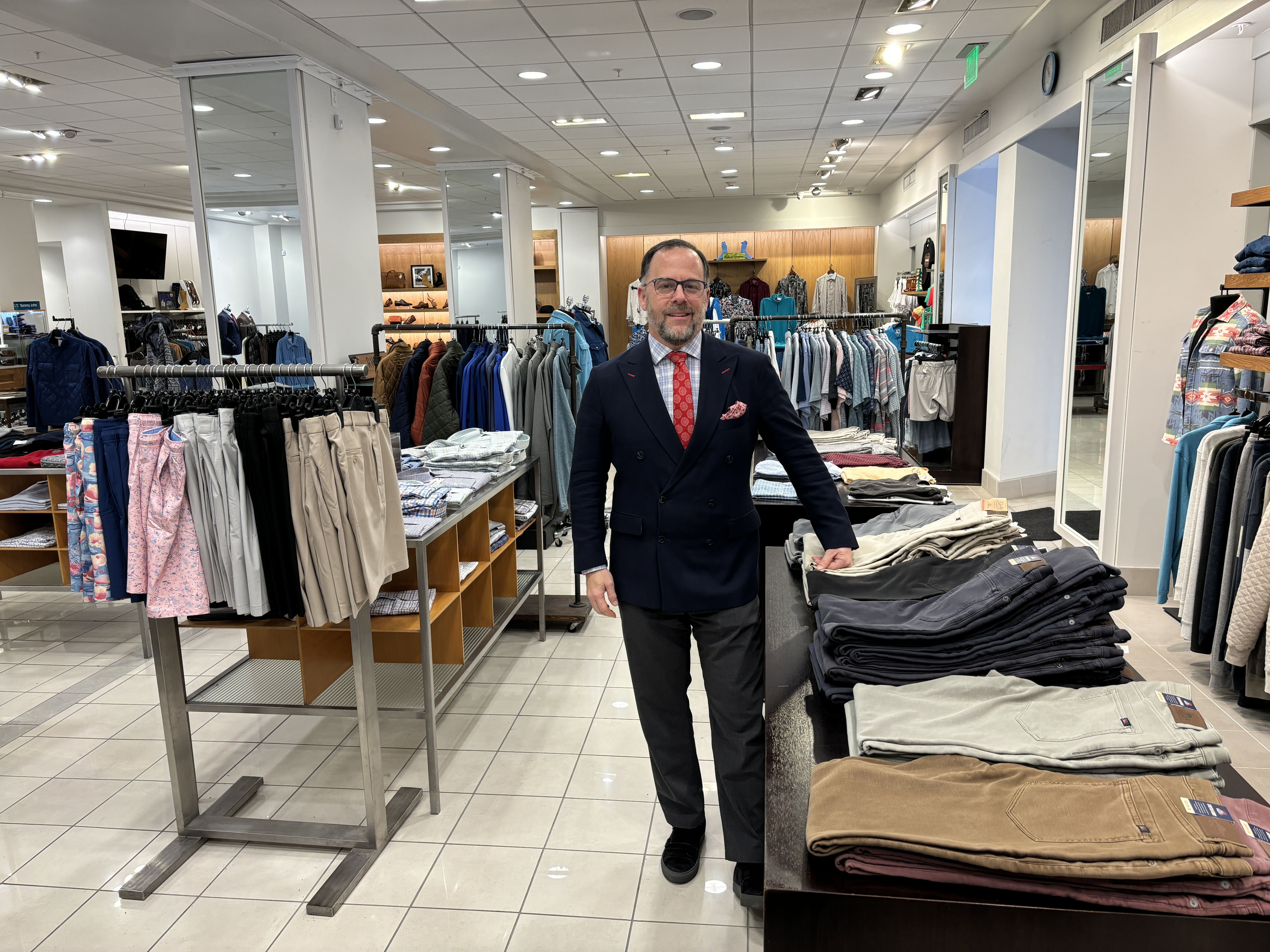 Kenny Rubenstein poses for a photo inside the Rubensteins store. Men's clothing is seen on racks and folded on tables around him.