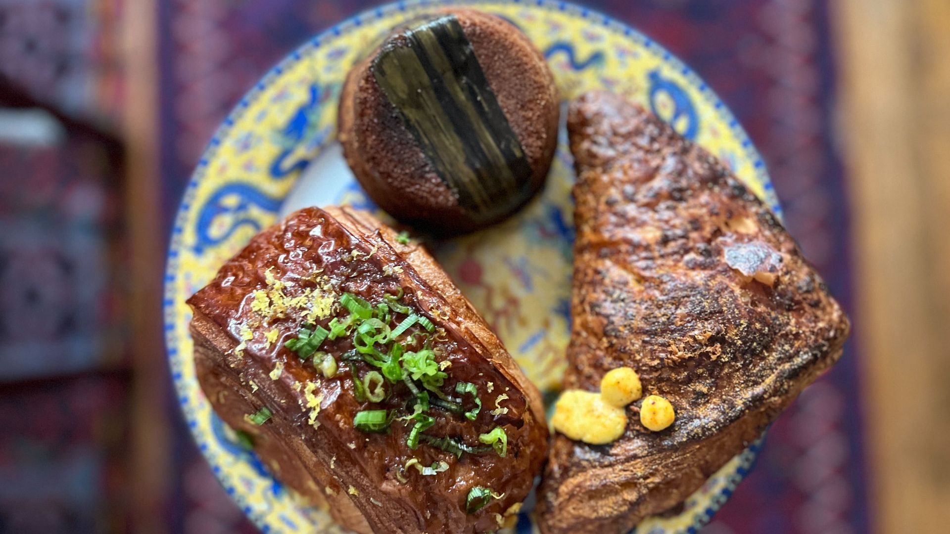 Plate with three baked goods: a rectangular glazed loaf topped with green herbs, a round dark bread with black stripe, and a triangular pastry with a dollop of yellow spread.