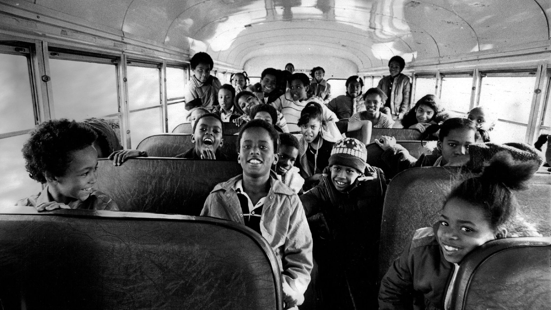Multiple children smile while seated inside a school bus, which has seats with a leather-like texture, and the bus' roof can be seen curving above the students. There are several windows on both sides. 