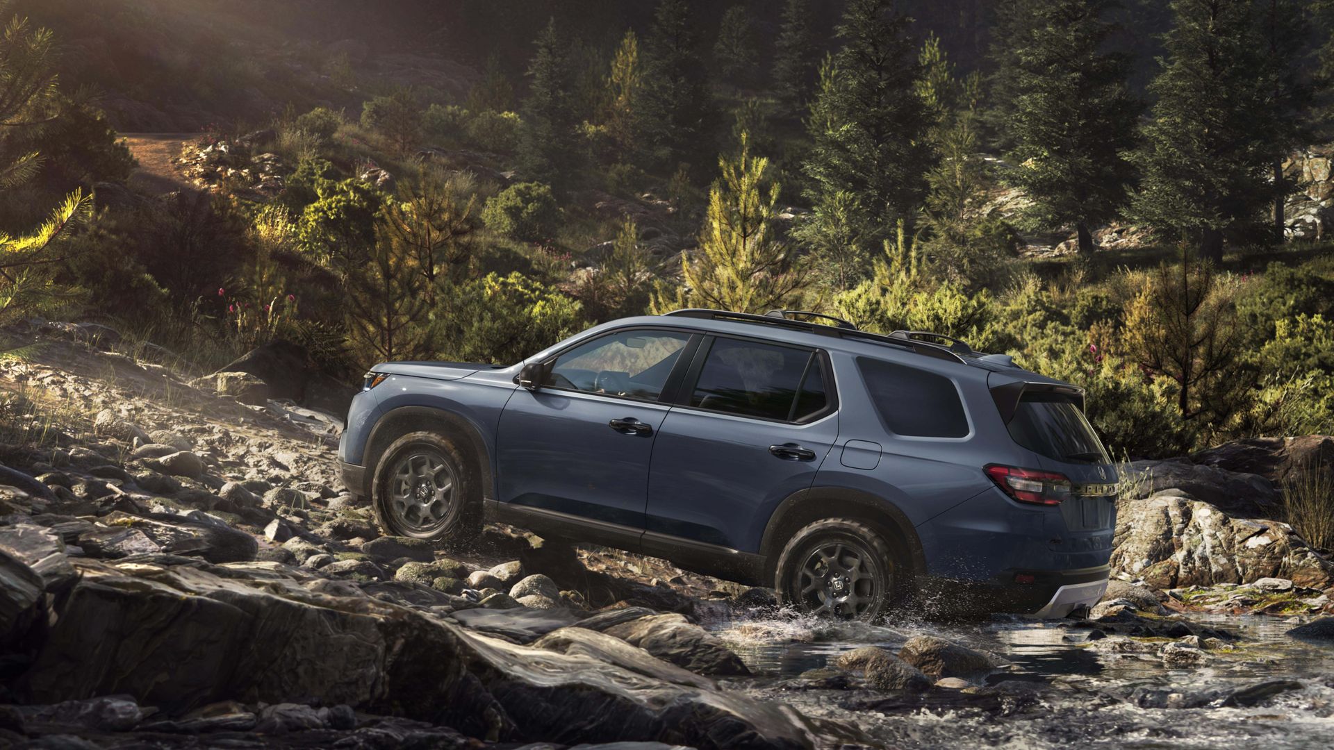 Side view of a blue Honda Pilot TrailSport SUV with roof rails climbing up a rocky riverbank, with a dense evergreen forest in the background and warm sunlight from the left.