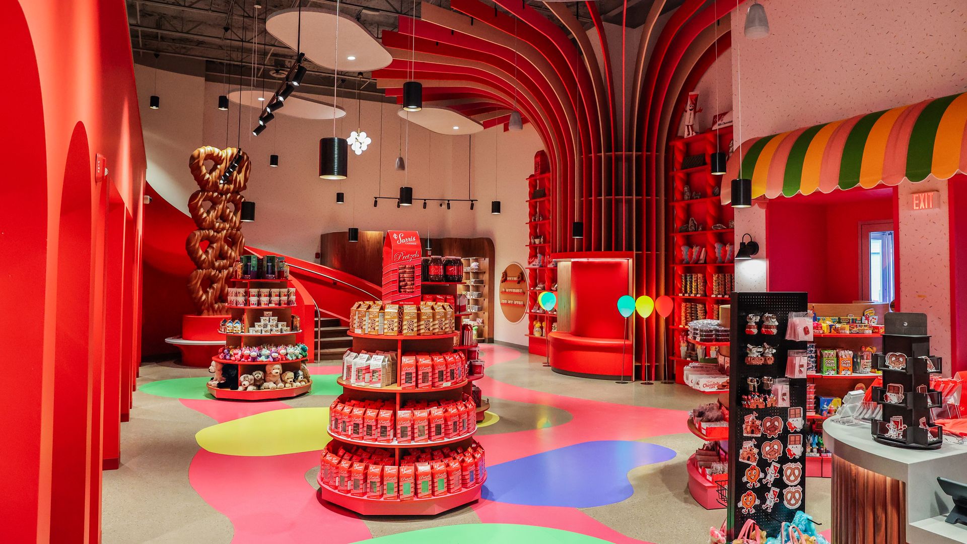 Bright candy shop interior with red walls, curved colorful floor patterns, circular displays, a large pretzel sculpture on the left, red arches, and a rainbow striped awning on the right.