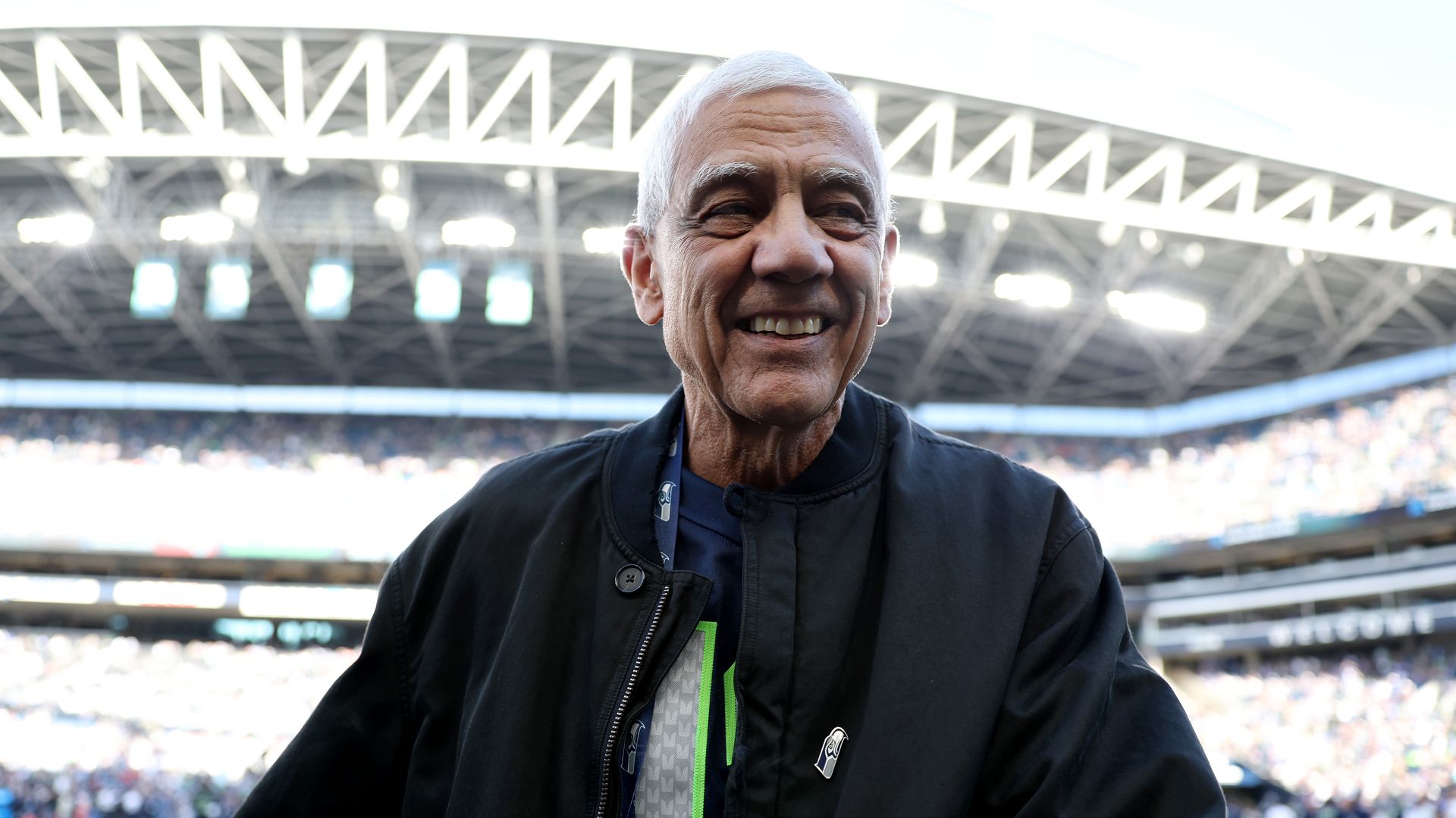 An older man with gray hair smiles at a stadium, wearing a black jacket. Bright stadium lights and a white truss roof loom behind, with a blurred crowd.