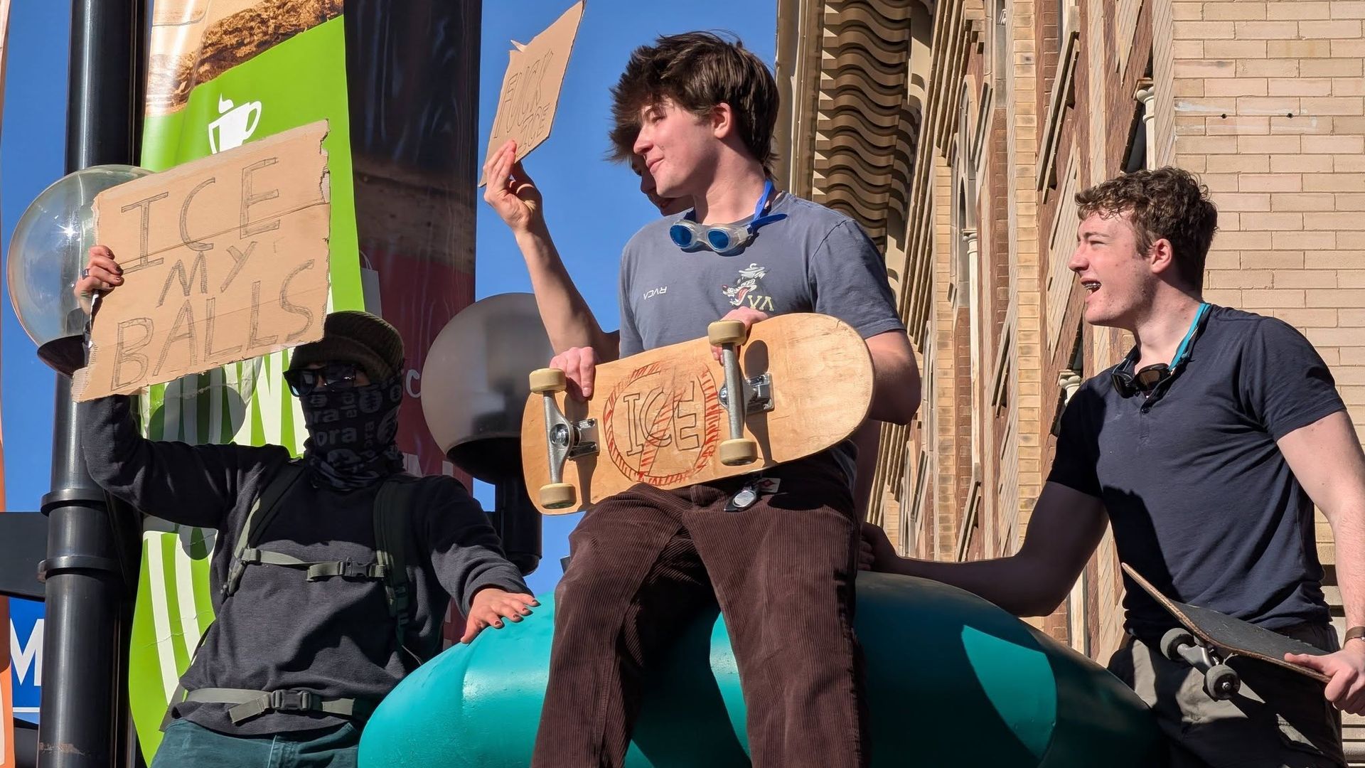 Three young men with skateboards stand on a public structure; one holds a cardboard sign reading "ICE MY BALLS," another holds a similar sign, and the third laughs, all under a clear blue sky.