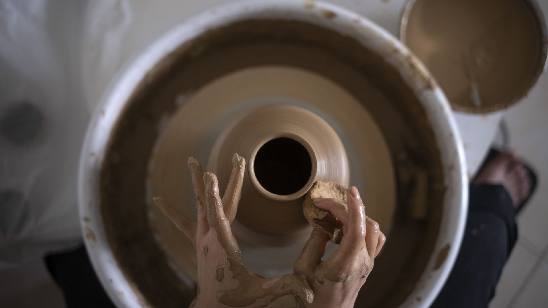 Top view of hands shaping a pottery vase on a spinning wheel, with clay-covered fingers and a sponge smoothing the surface.