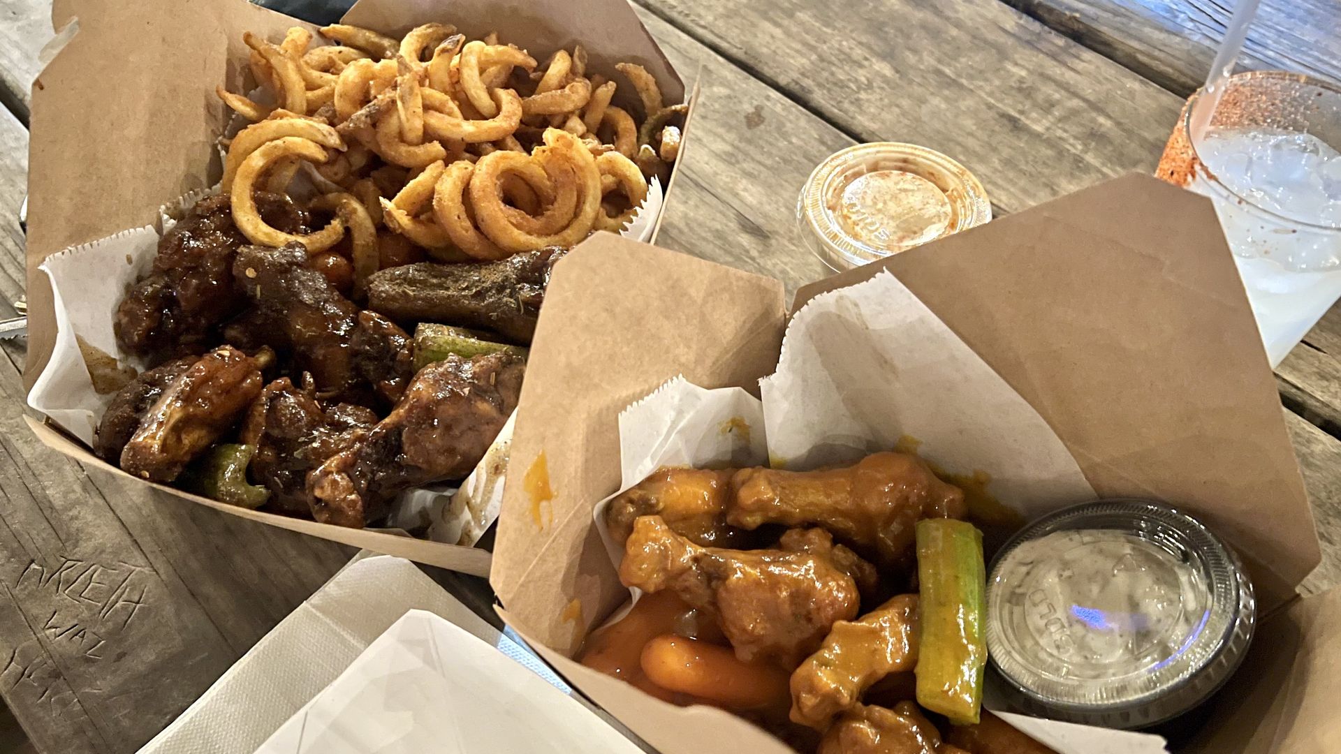 Top-down view of a wooden table with three open brown takeout boxes: golden onion rings, saucy fried chicken wings, and orange chicken bites with celery. A small sauce cup and an iced drink sit nearby.