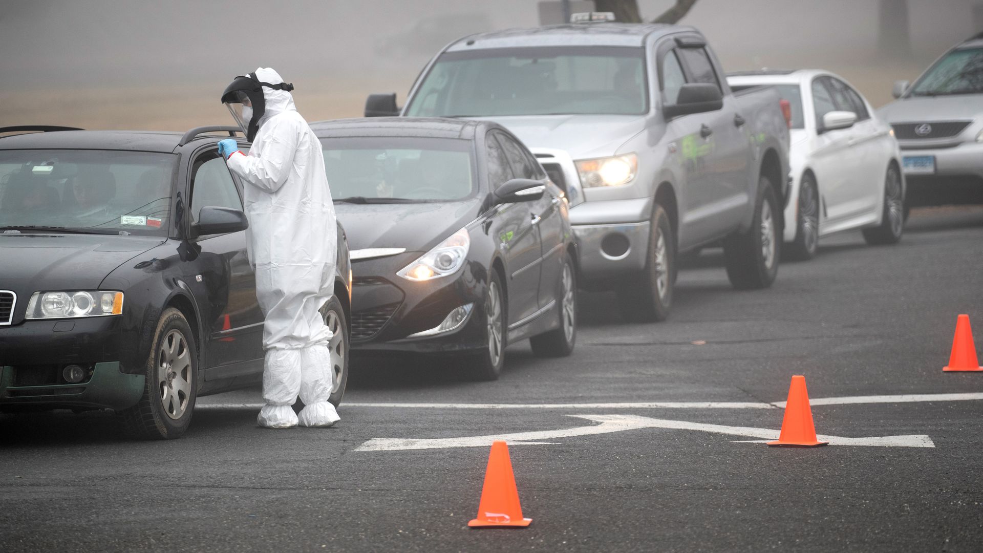 In this image, a worker stands in a protective medical suit next to a line of cars