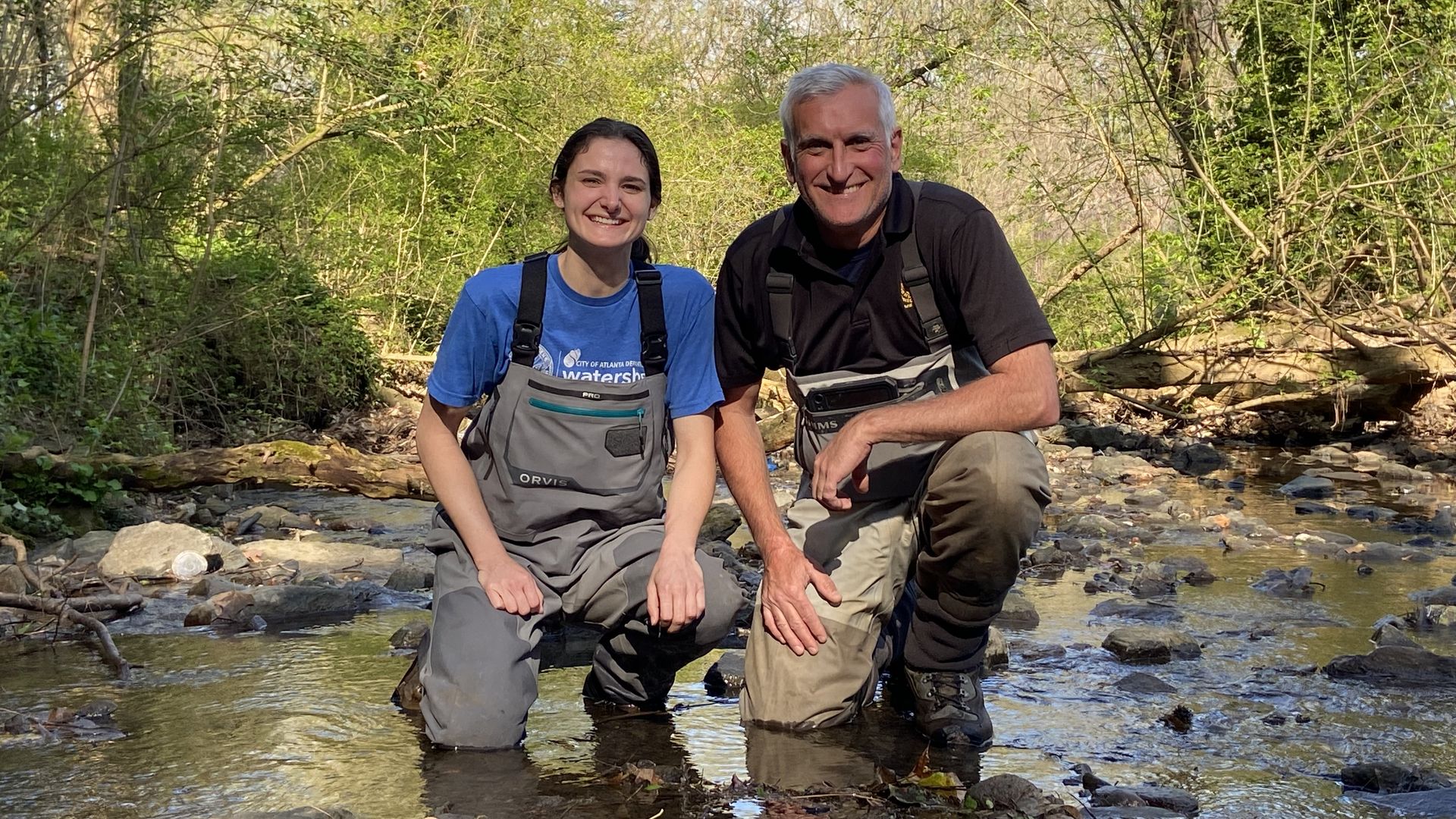 Two people wearing waders take a knee in a stream in the woods