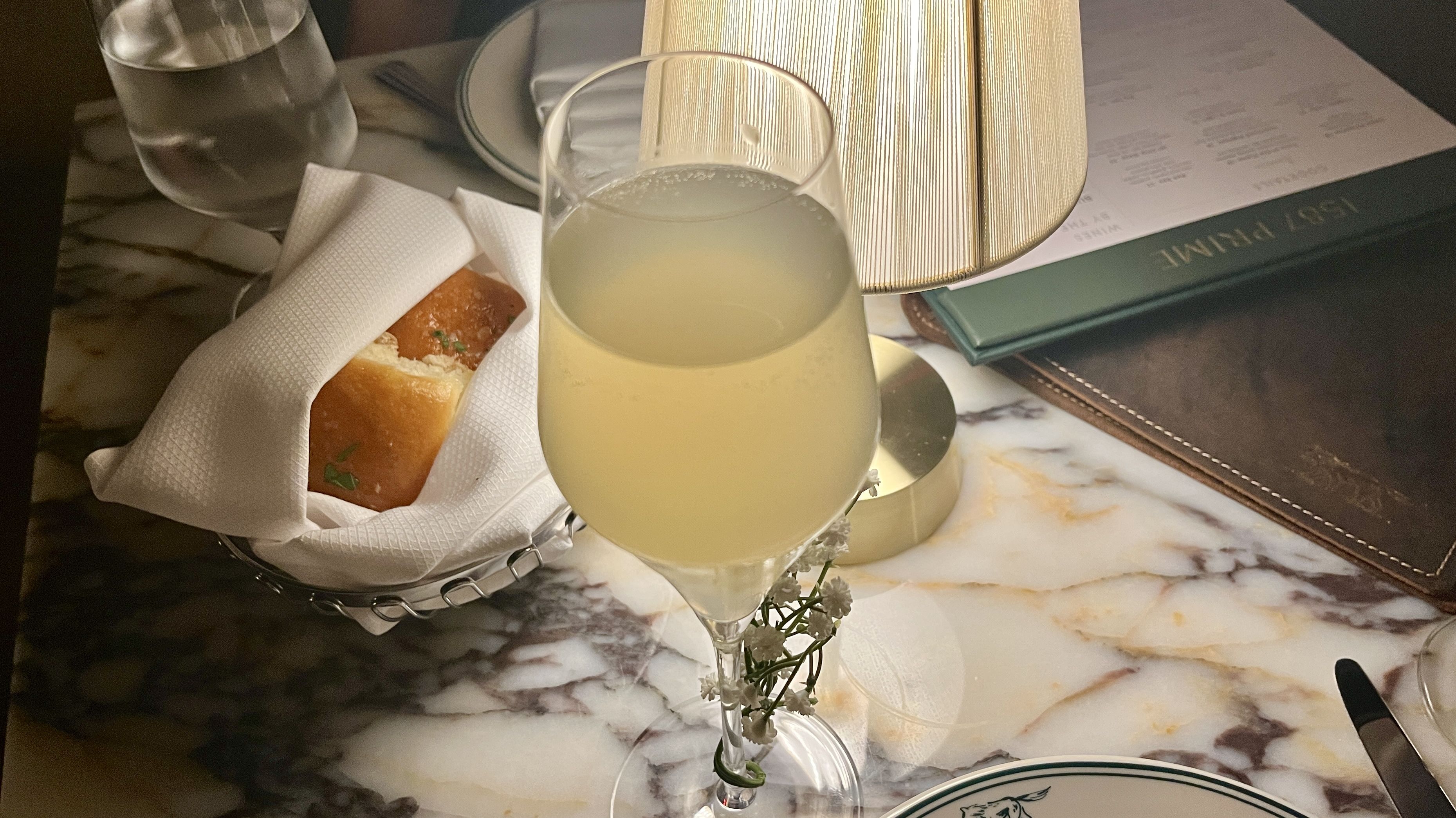 A glass of pale yellow sparkling drink with small white flowers tied to the stem, a bread roll wrapped in a white napkin, a water glass, and a menu on a marble table lit by a small lamp.