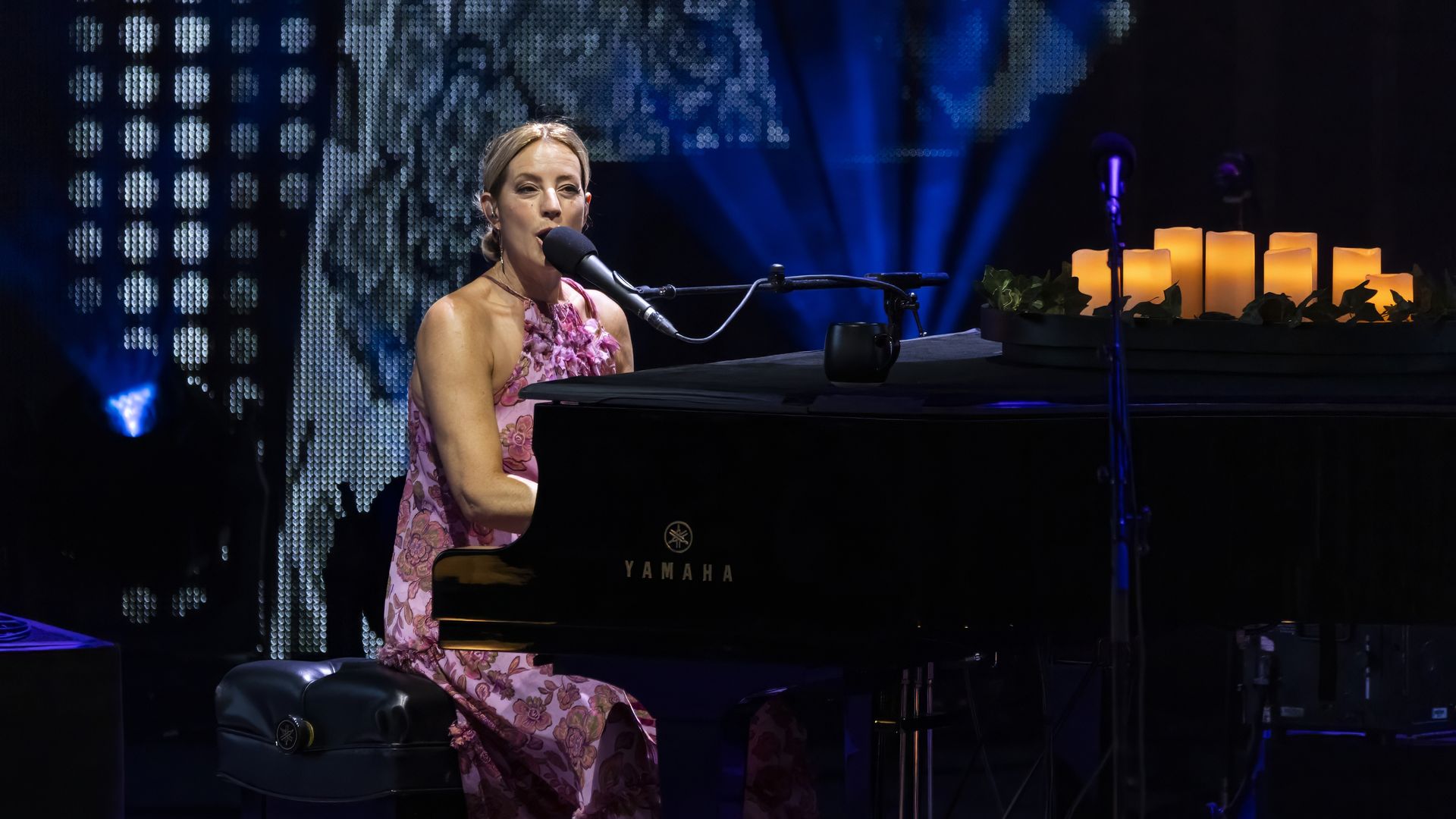 Sarah McLachlan sings into a mic while playing the piano. Candles sit on the piano.