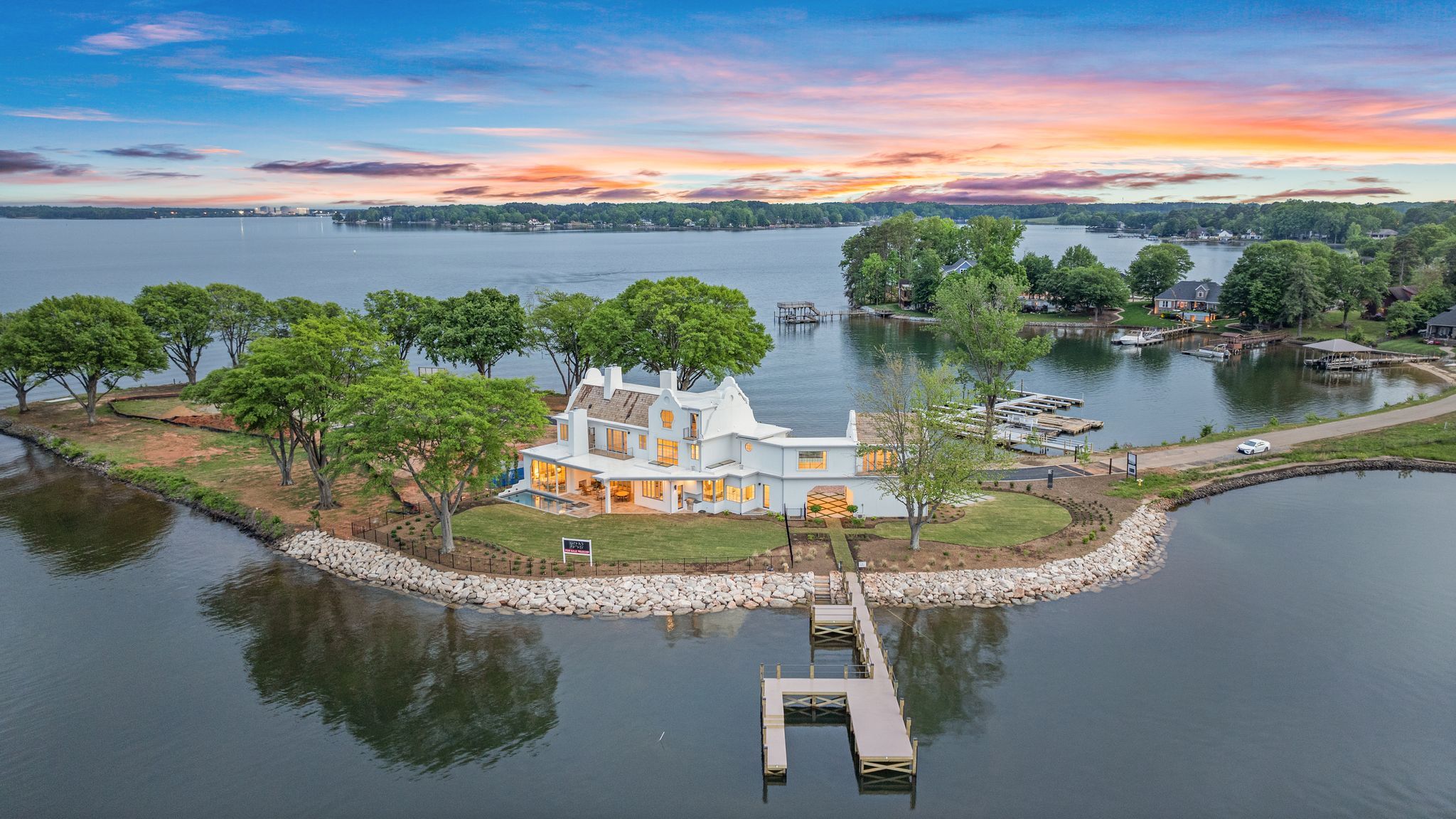 A large white house with illuminated windows on a peninsula, surrounded by water and green trees, with a dock extending into the lake under a colorful sunset sky.