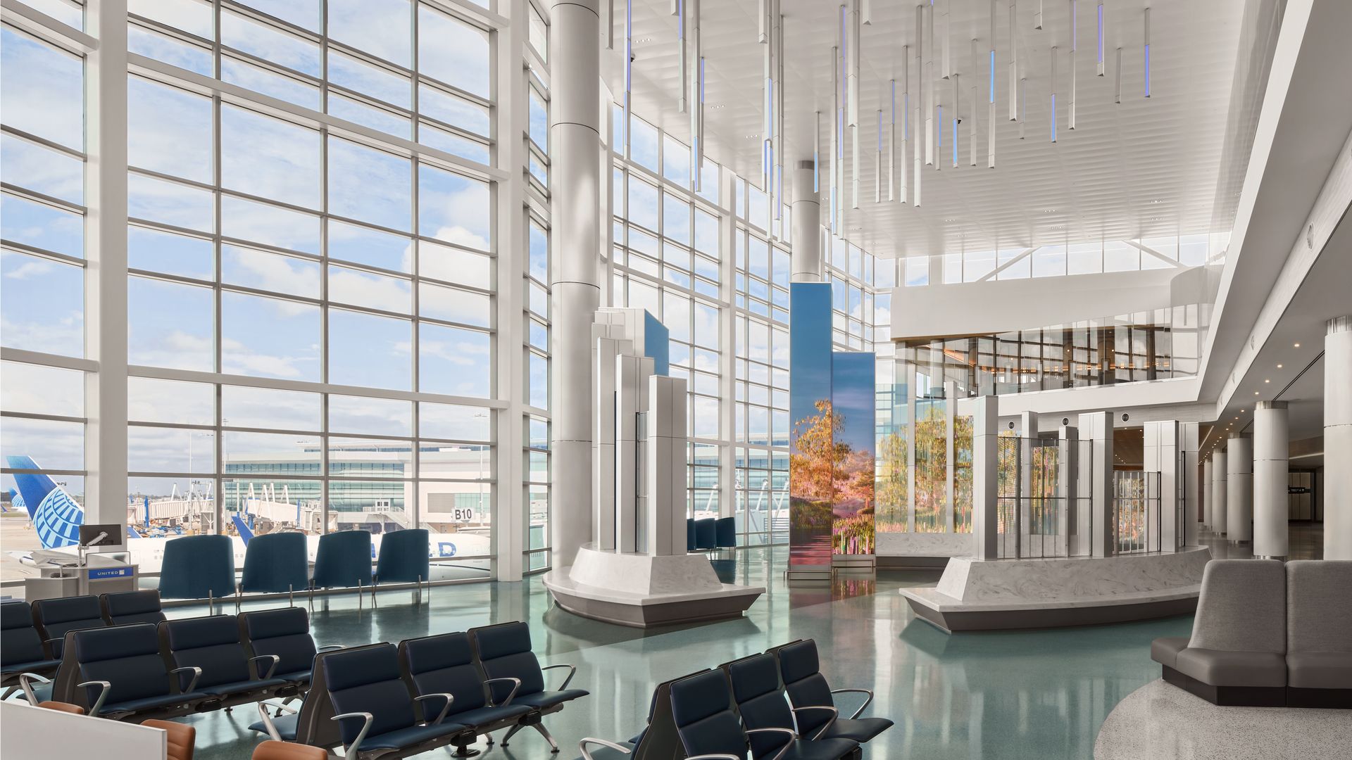 Bright airport terminal lounge with tall glass walls, blue and orange seats, white pillars, and a modern ceiling with hanging light tubes; planes visible outside.