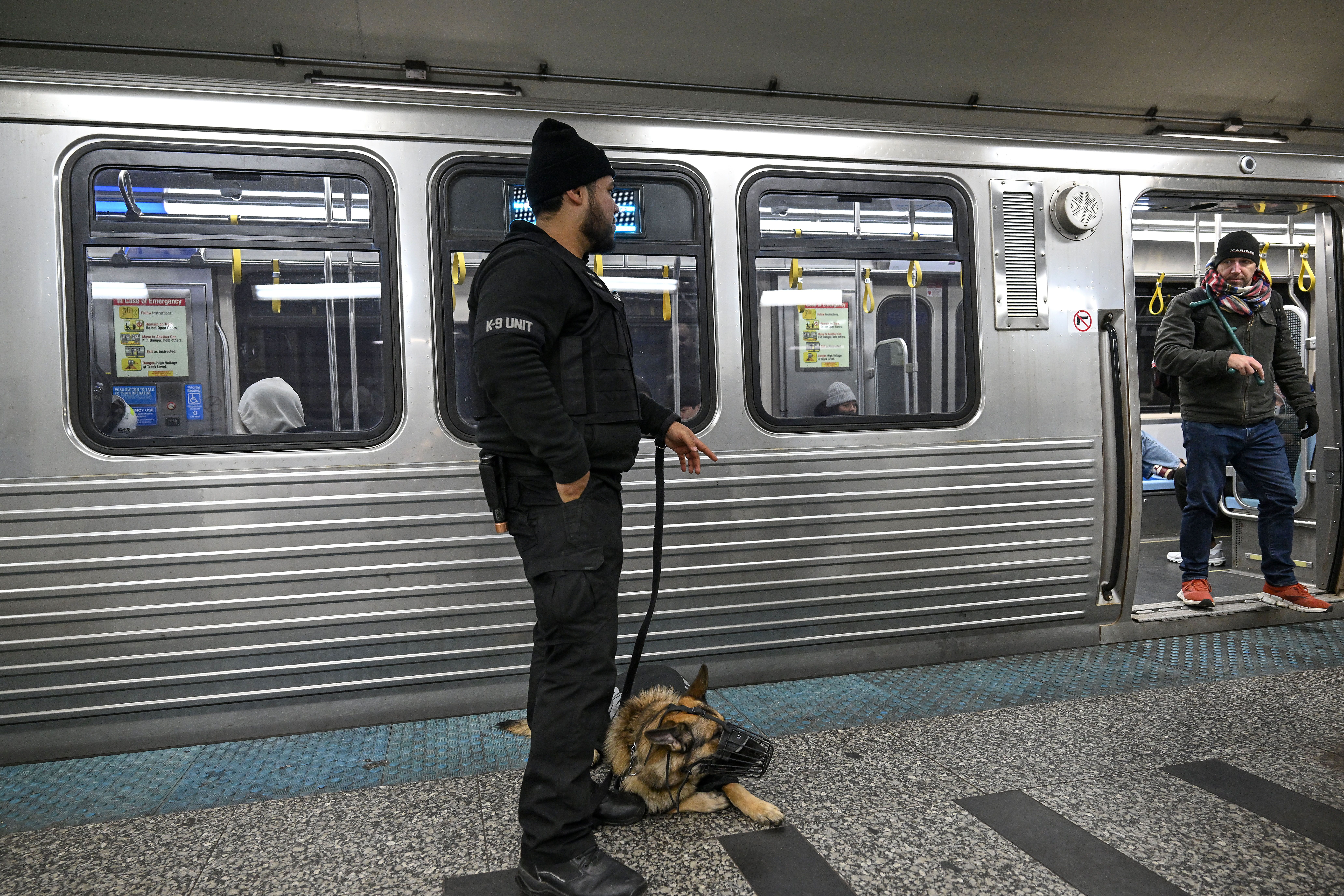 A K-9 unit officer in black stands on a subway platform holding a leash of a muzzled German Shepherd dog. A man wearing a scarf and hat stands in the subway car doorway.