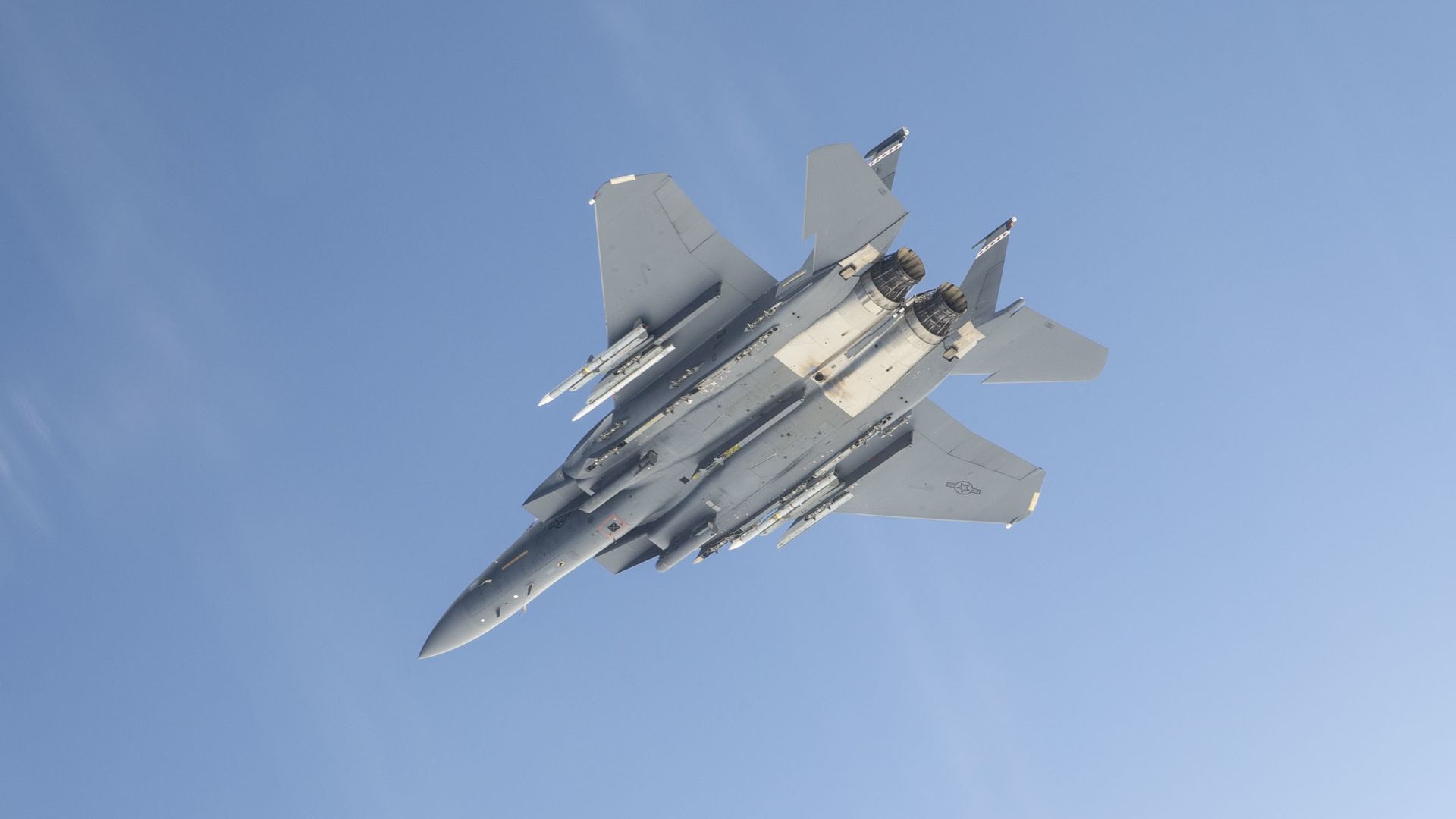 The belly of an F-15E, armed with advanced air-to-air missiles, is seen. The sky behind the aircraft is light blue.
