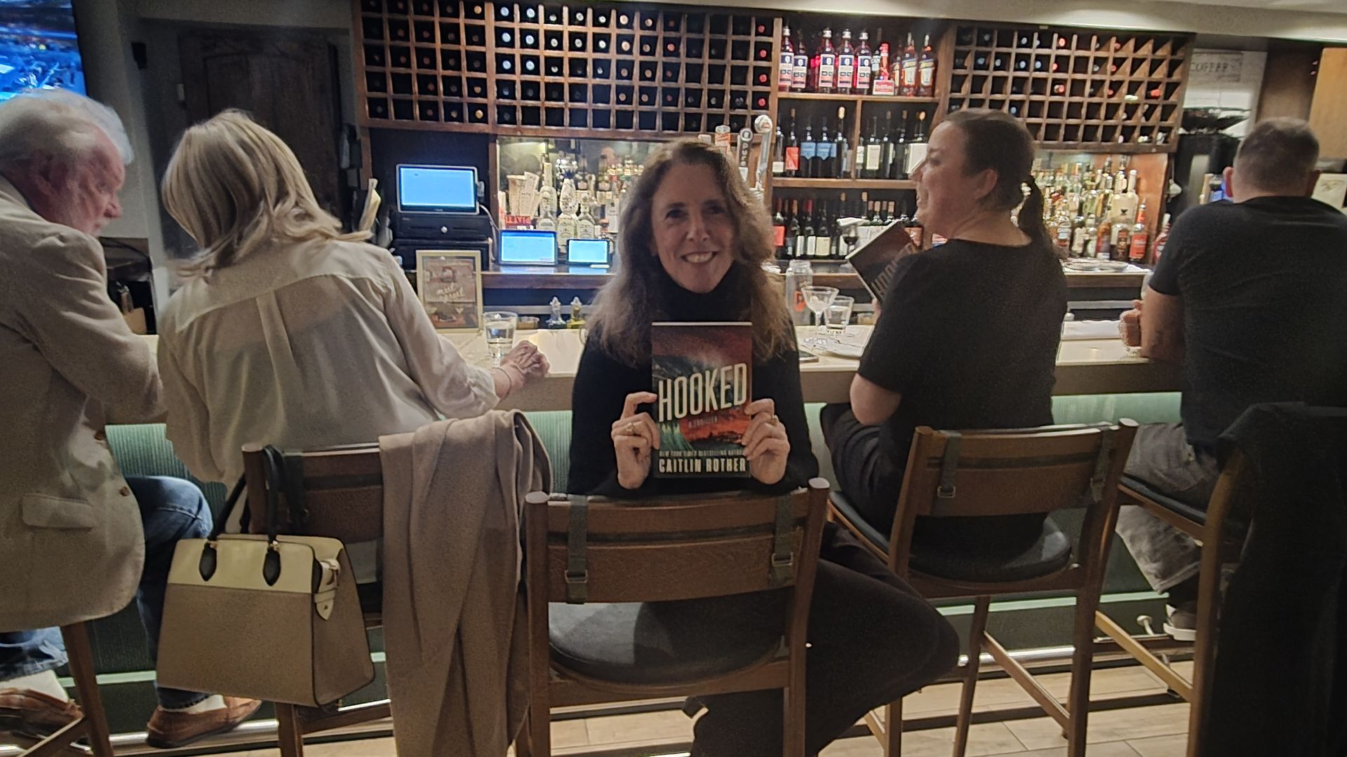 Smiling woman seated at a bar holding a book titled "Hooked" by Caitlin Rother, surrounded by four other people sitting on wooden chairs at the counter with bottles and wine racks in the background.