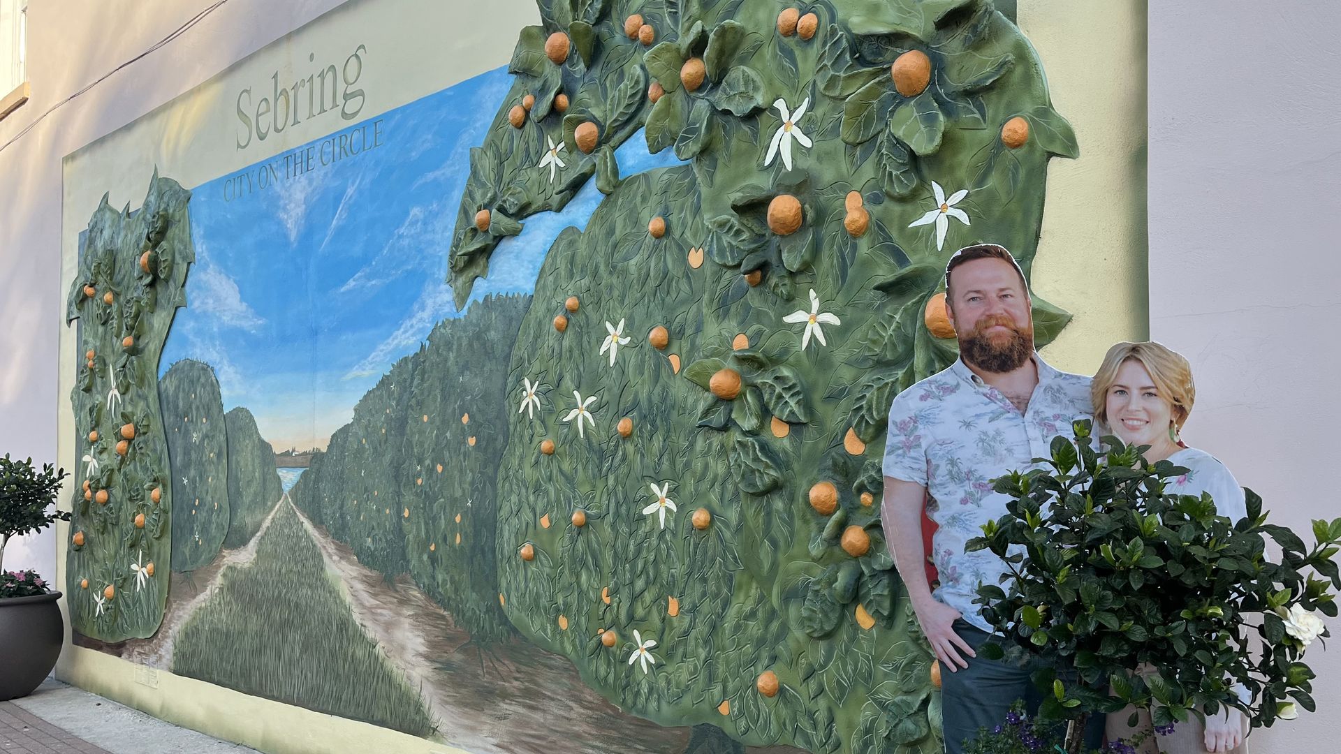 A cardboard cutout of a couple poking out from a bush beside a mural of an orange grove that says "Sebring, City on the Circle."