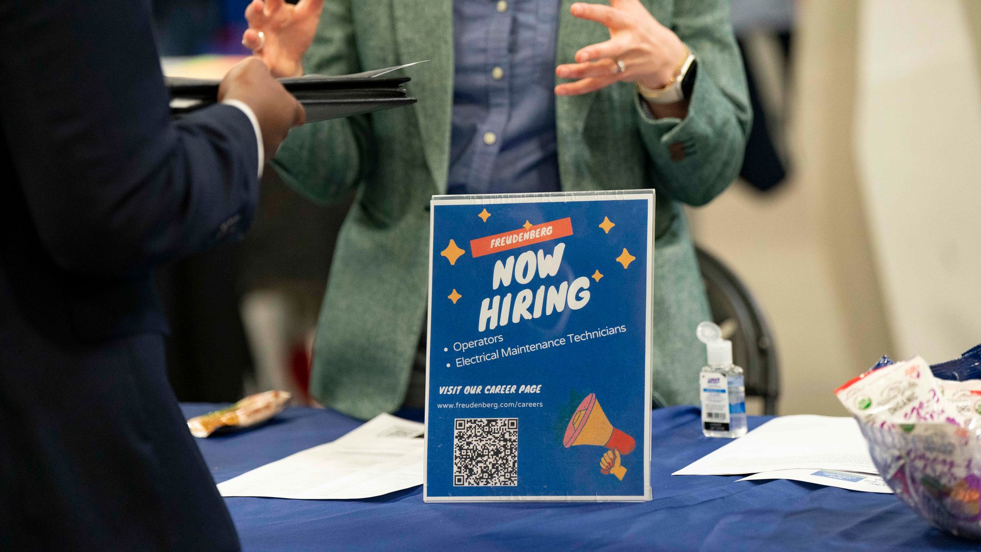 A person in a green blazer talks to another holding folders across a table with a blue sign reading "Now Hiring"