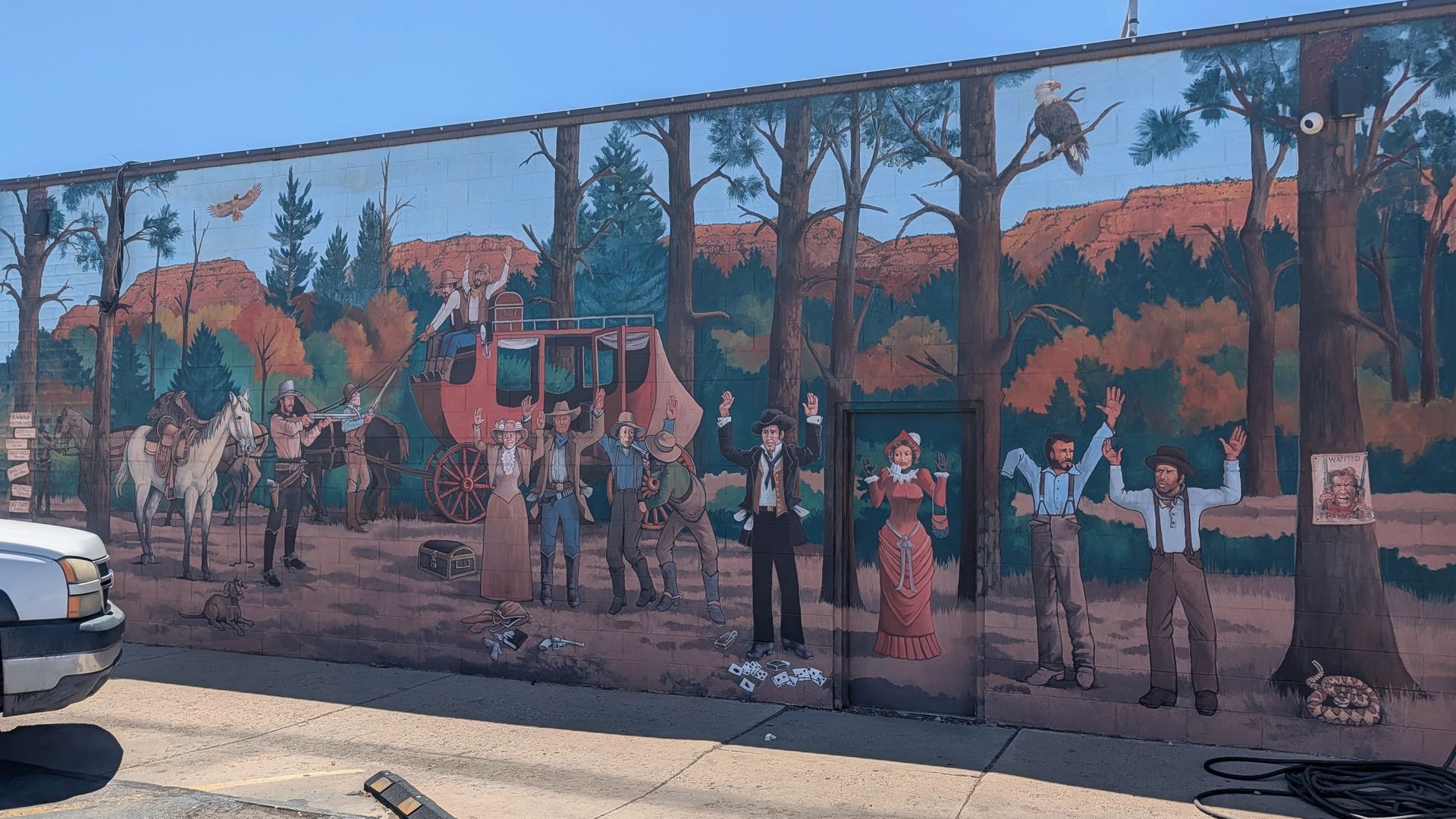 Wide mural of an Old West scene: cowboys around a red stagecoach, townspeople with raised arms, tall trees and orange desert hills, a Wanted poster on a tree, and a parked car in the foreground.