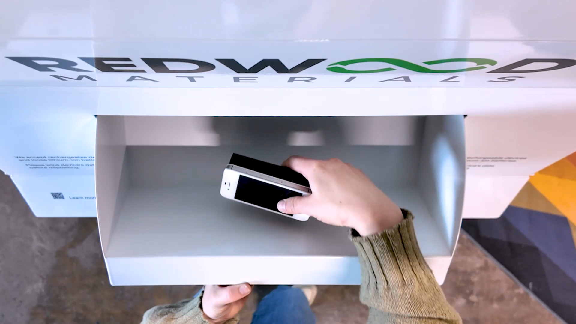 A person wearing a green sweater places an old white smartphone into a white recycling bin labeled "REDWOOD MATERIALS" with a green infinity symbol.