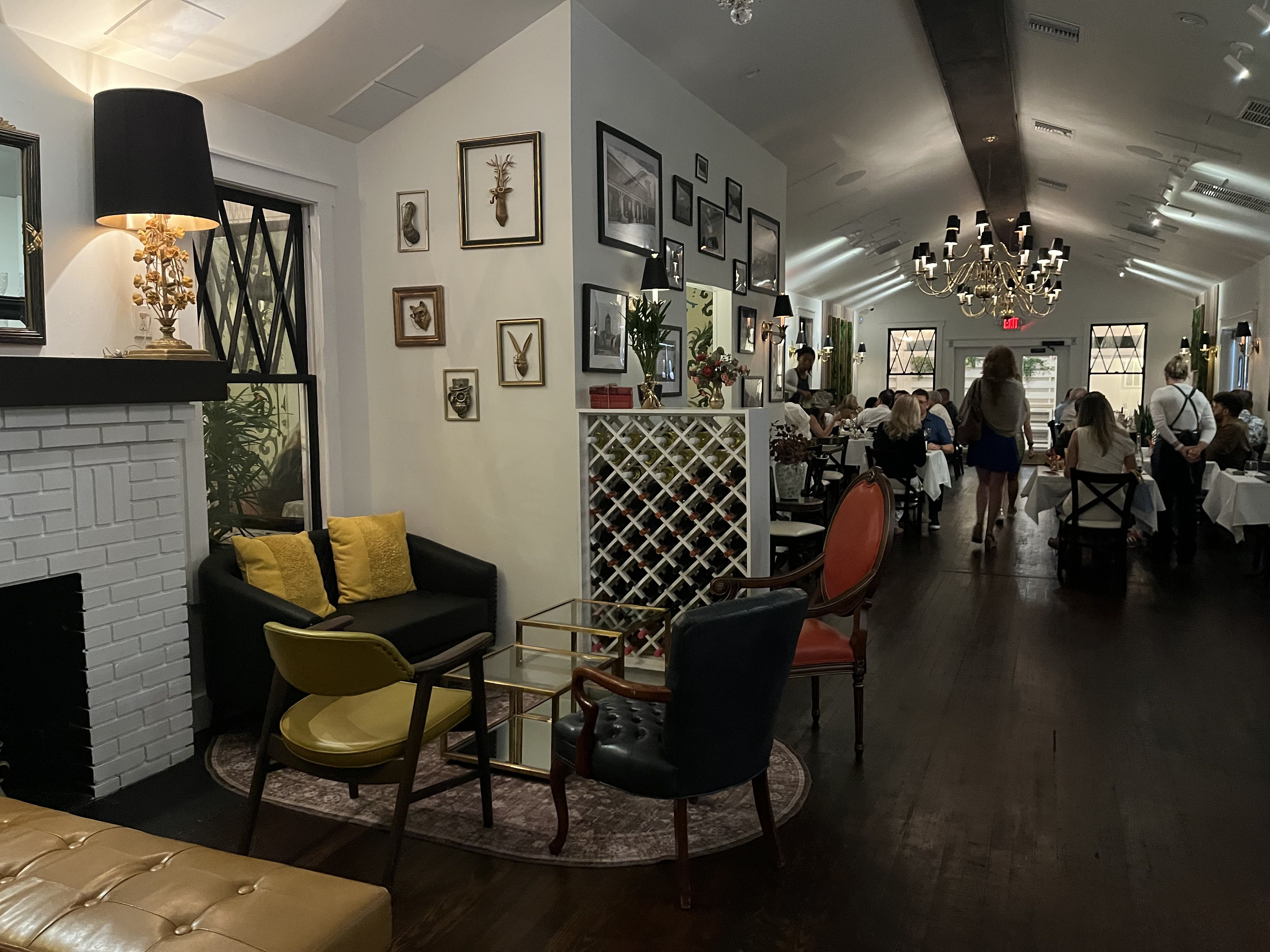 Elegant restaurant interior with white walls, framed art, and a large chandelier. Diners sit at white-clothed tables along a long corridor; black lamps, lattice dividers, and colorful seating.