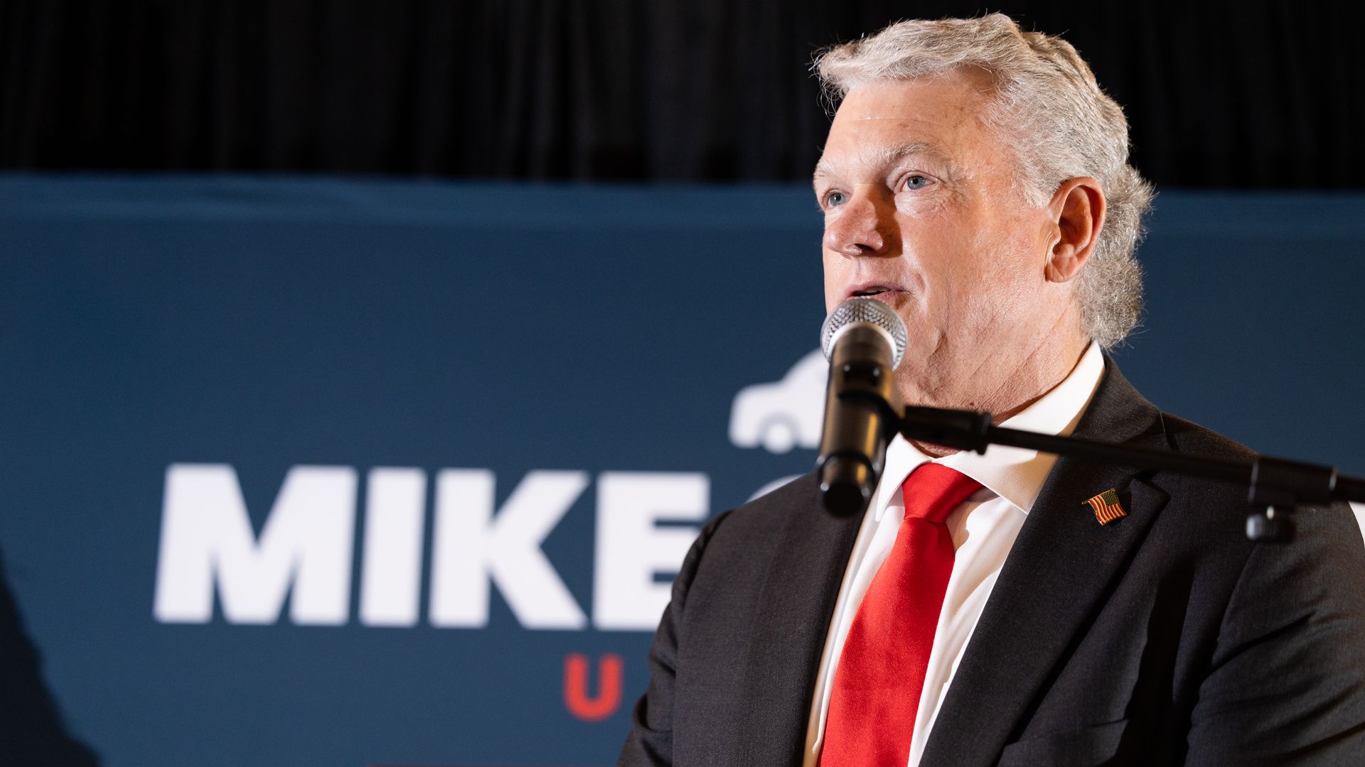 A man wearing a black sports jacket with an American lapel pin, red tie and white collared shirt speaks into a microphone. The words "MIKE" appear in white on a blue background. 