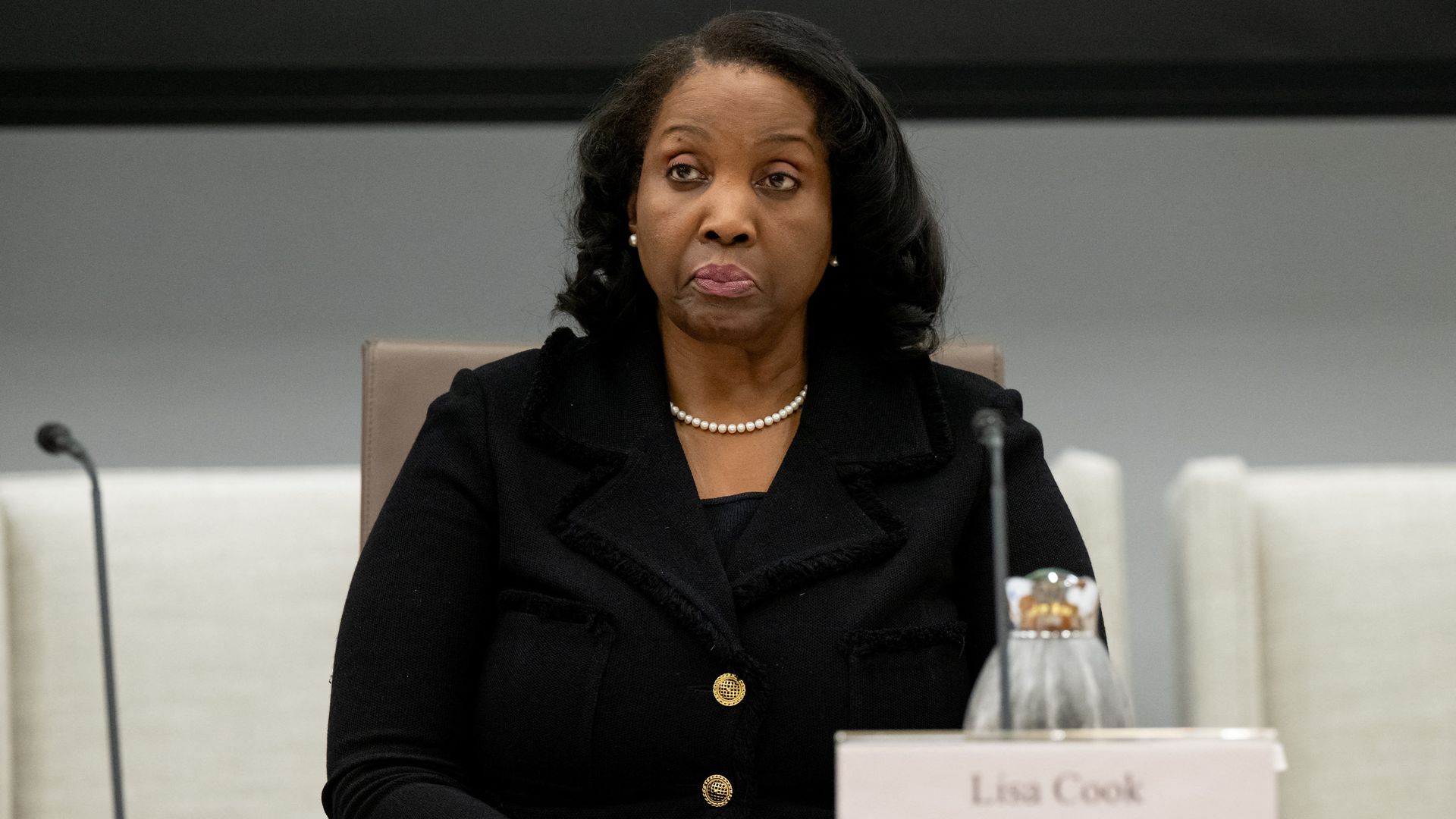 A woman wearing a black jacket and pearl necklace is seated at a conference table with a nameplate reading "Lisa Cook Governor" in front of her.
