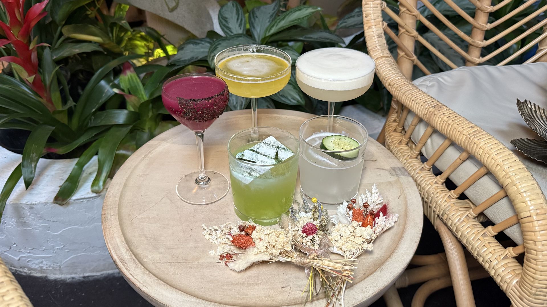 Round wooden table with five cocktails: pink raspberry-rimmed, yellow citrus, white foam, green with ice, and pale gray cucumber. A wicker chair and lush tropical plants fill the background.
