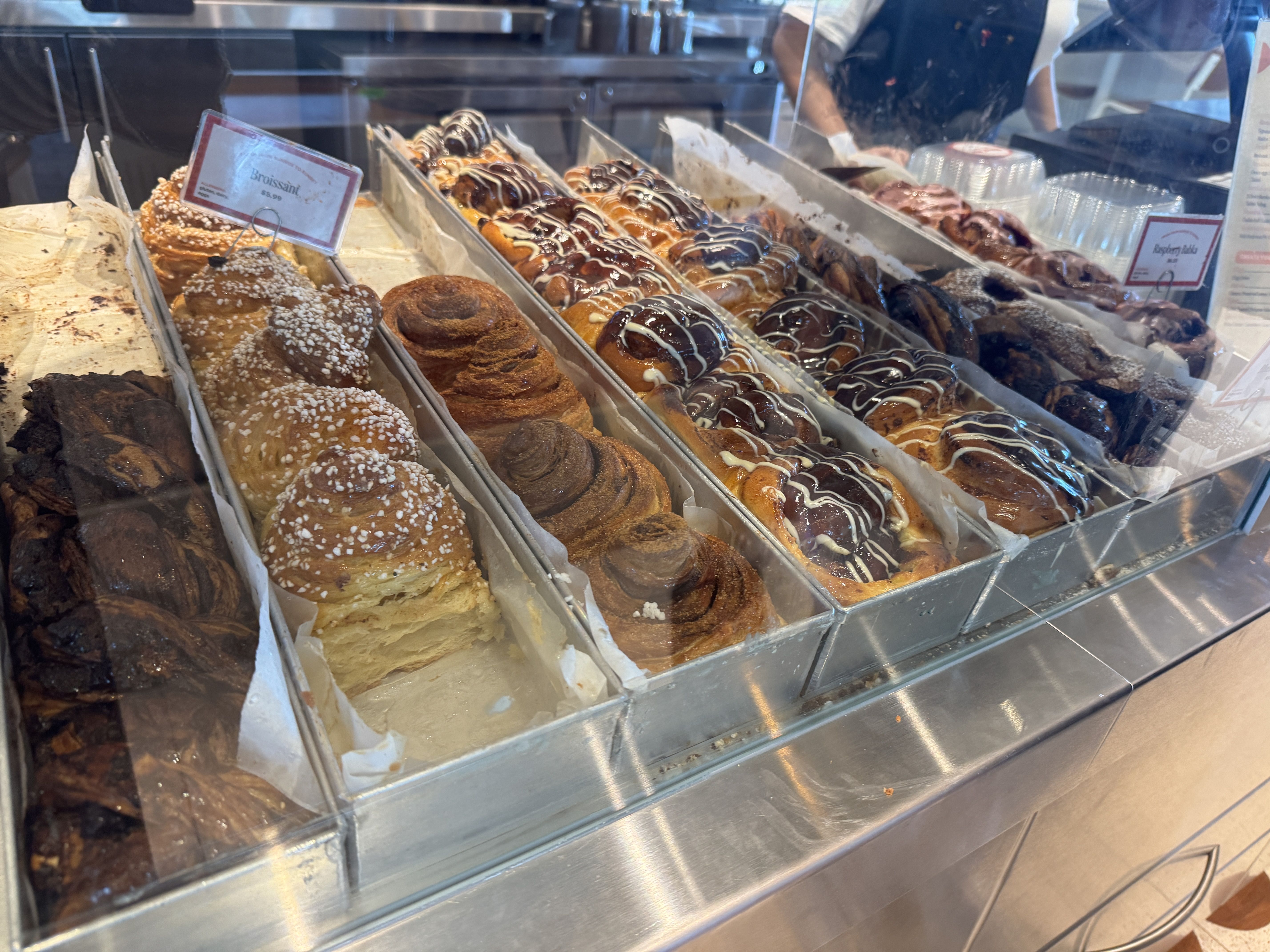 Bakery display case with an assortment of pastries: babka broissant, cinnamon babka, chocolate-drizzled babka, and swirled iced babka arranged in metal trays behind glass.