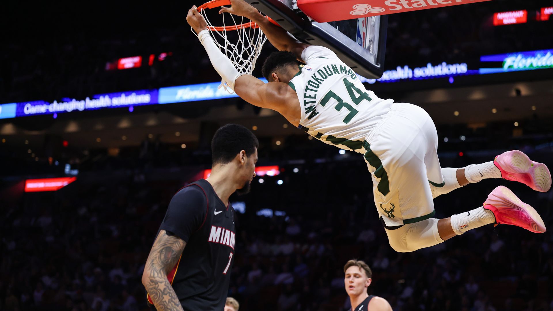 A man in a white Milwaukee Bucks jersey reverse-dunks a basketball as two Miami Heat players in black jerseys look at each other, befuddled.