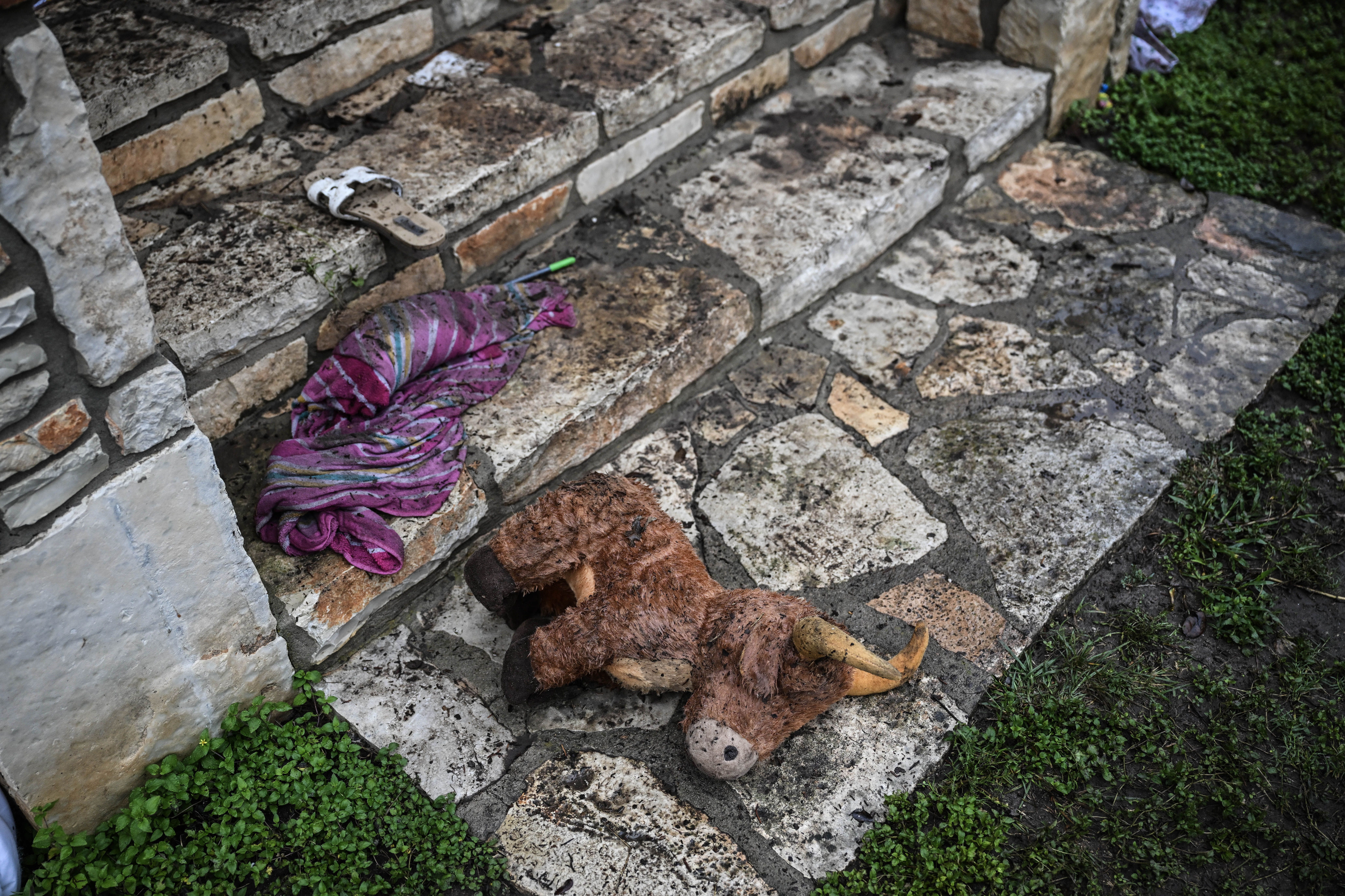 Dirty stuffed bull toy lies on stone steps outside, next to a muddy purple cloth and a white sandal, with grass growing beside the stone path.